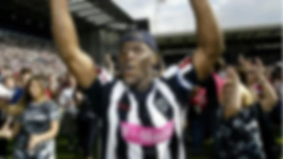 Blurred image of people celebrating at a sports event, with a focus on a person in a black and white striped jersey raising their arms.