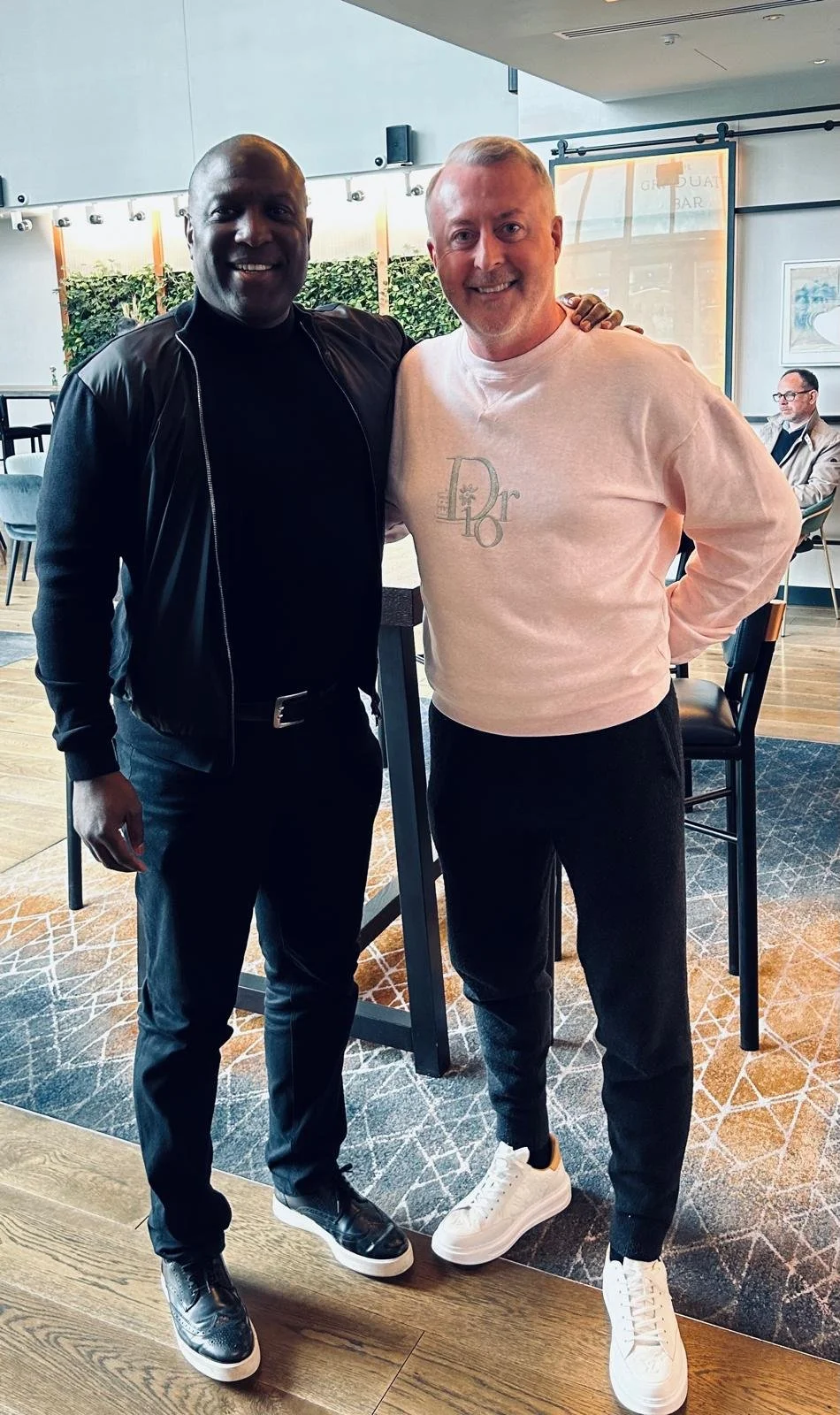 Kevin Campbell and a good friend smiling and posing for a photo indoors, one wearing a black jacket and the other a light-colored sweater. Background includes a seating area and a person sitting at a table.