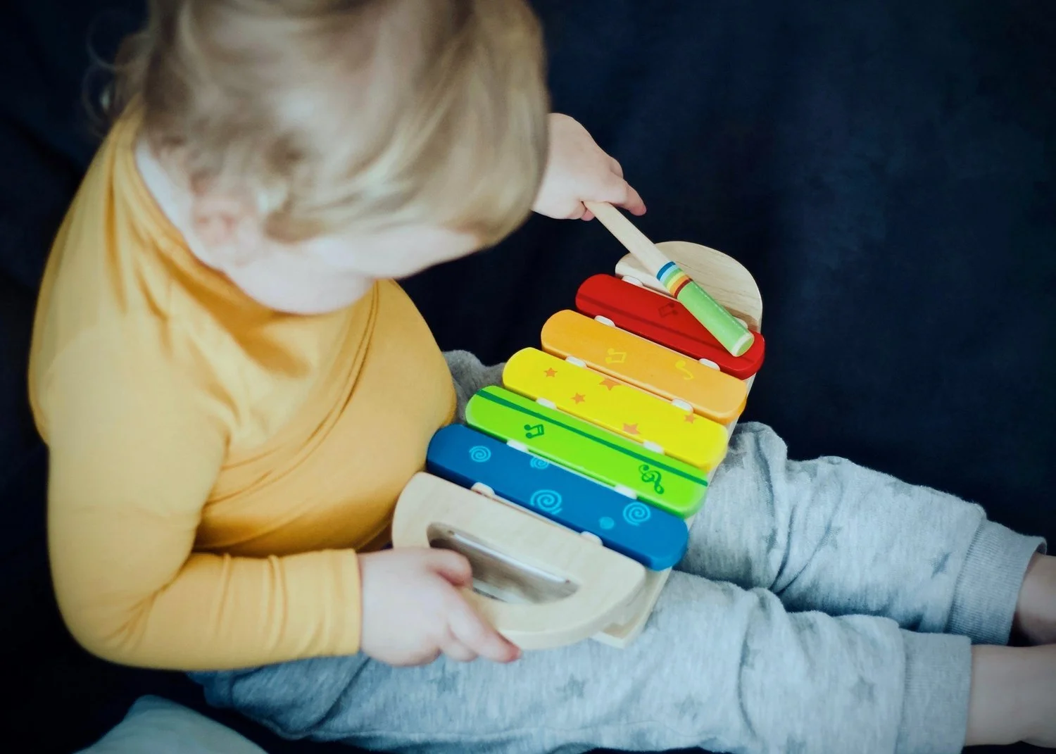 A young child with blond hair, wearing a yellow long-sleeve shirt, sitting on a dark surface, playing a colorful wooden xylophone with a green mallet.