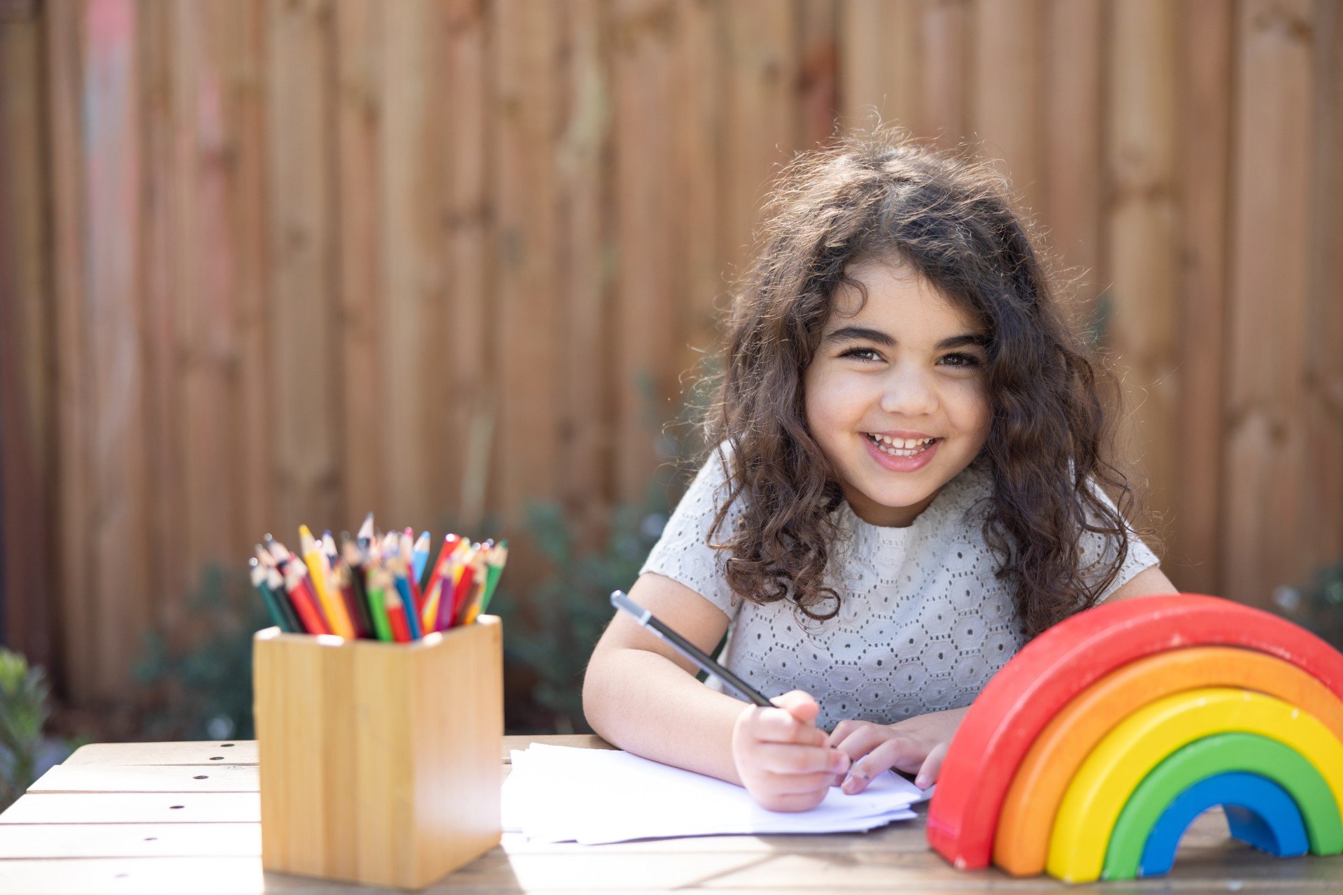 A young girl with curly brown hair smiling and drawing at a wooden table outdoors, with a rainbow stacked toy and a container of colored pencils nearby.