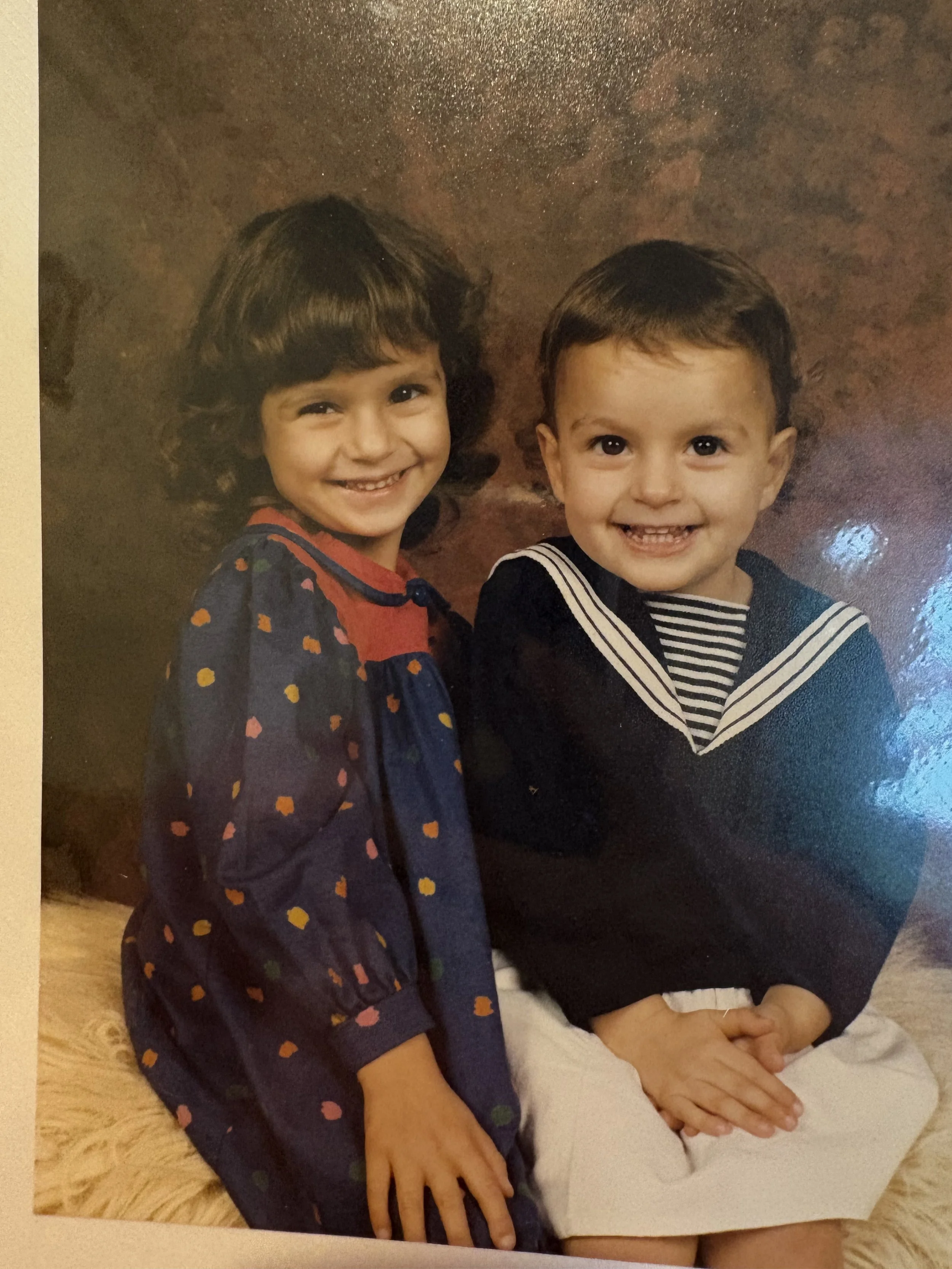 Two young children, a girl with dark curly hair wearing a colorful dress and a boy with short dark hair in a sailor outfit, smiling and sitting together against a brown textured background.