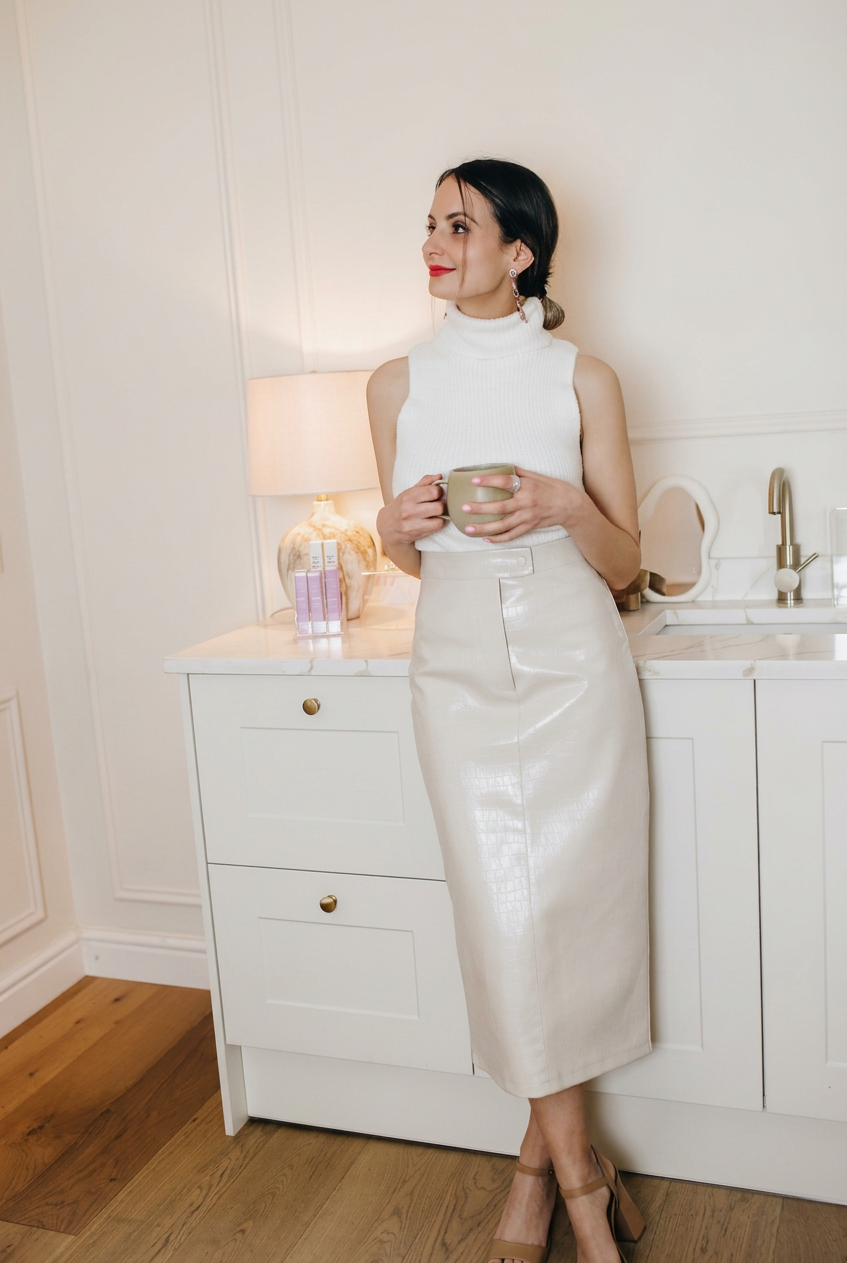 Woman in a white sleeveless turtleneck top and a shiny beige midi skirt standing in a kitchen, holding a beige mug, with a lamp and decorative items on the counter behind her.