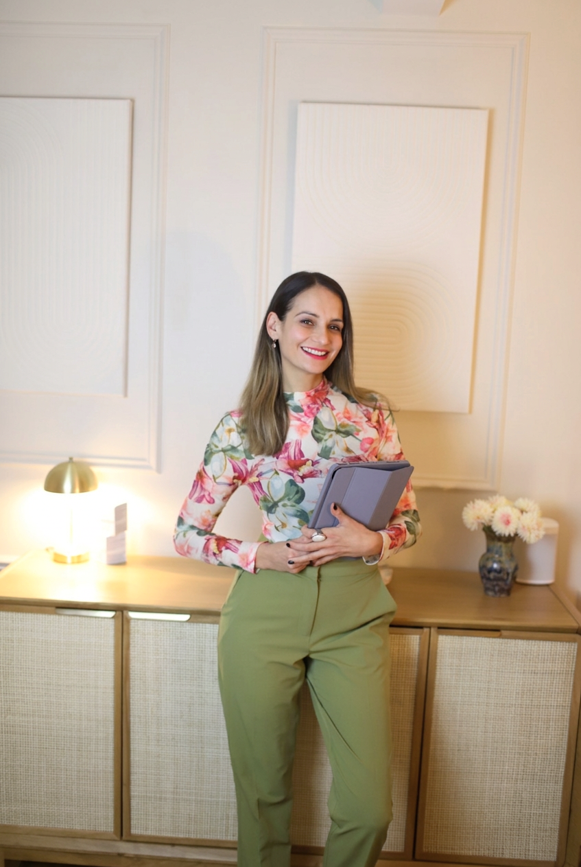 A woman with shoulder-length brown hair, smiling, wearing a floral blouse and green pants, holding a tablet, standing in a modern, well-lit room with beige walls, a wooden sideboard, a small lamp, and a vase of flowers.