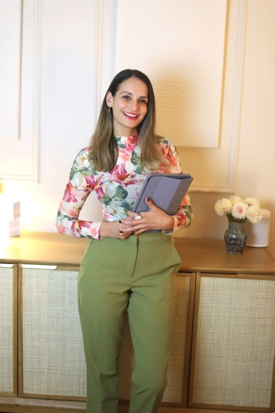 A smiling woman with shoulder-length brown hair, wearing a colorful floral long-sleeve top and green pants, holding a closed tablet computer, standing in a warm-toned room with a wooden cabinet, a blue vase with white flowers, and a lit lamp.