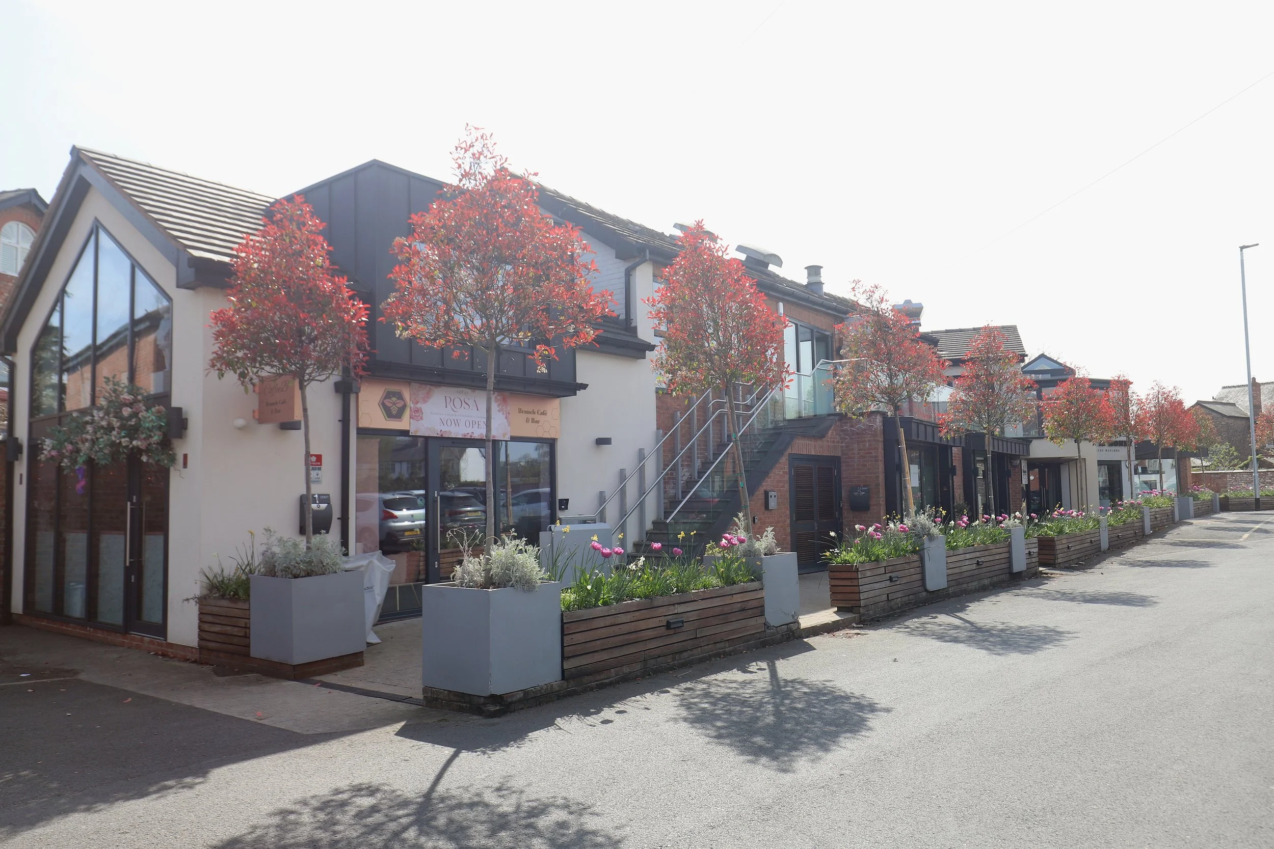 Row of modern commercial buildings with large glass windows, decorative trees, and flower planters on the sidewalk, under a partly cloudy sky.
