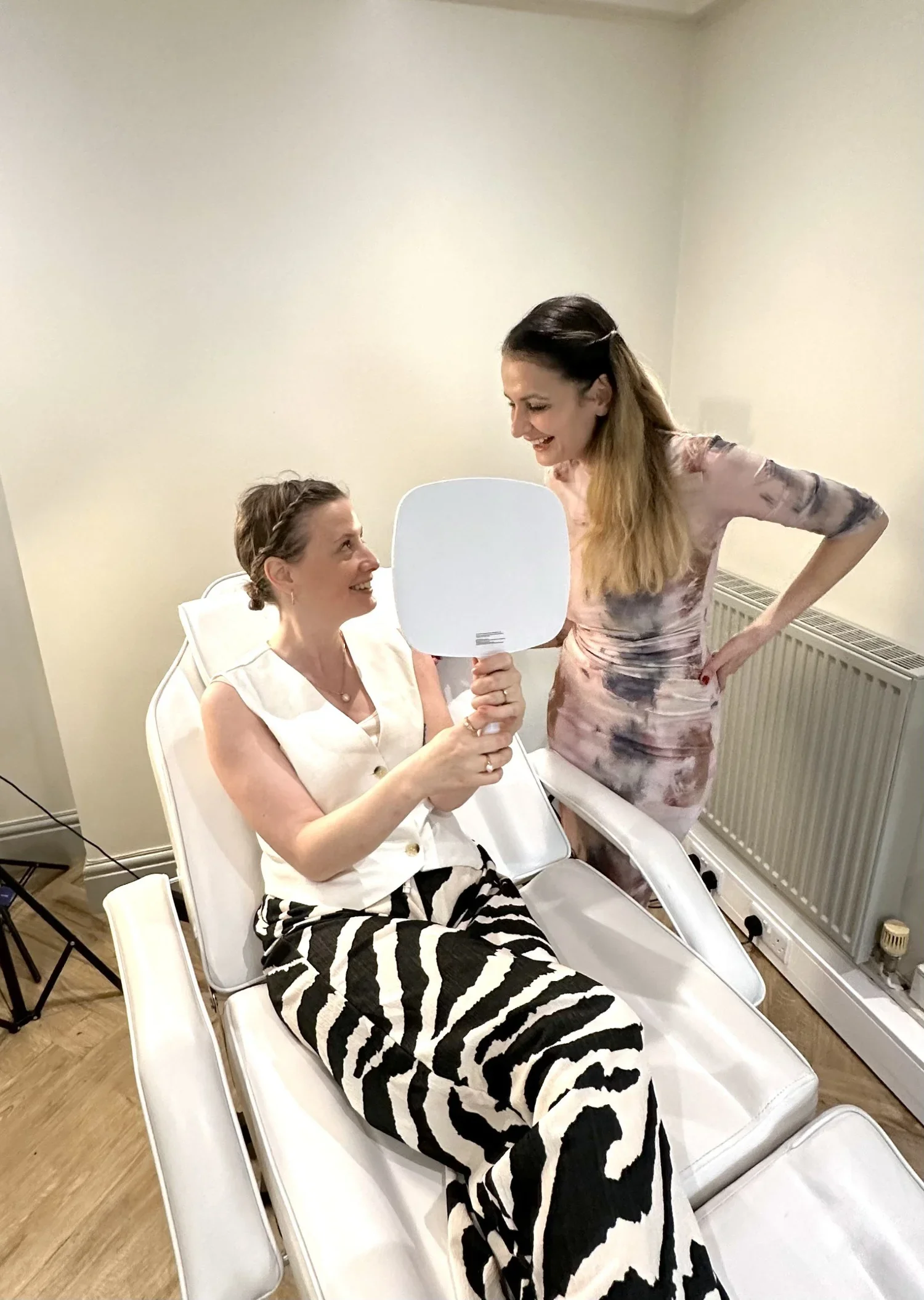 A woman sitting on a white chair in a medical setting, holding a handheld mirror and smiling at another woman standing nearby, who is smiling back and leaning forward. The standing woman has long hair and is wearing a floral dress. The sitting woman has short hair and is dressed in a white top and zebra-striped pants.