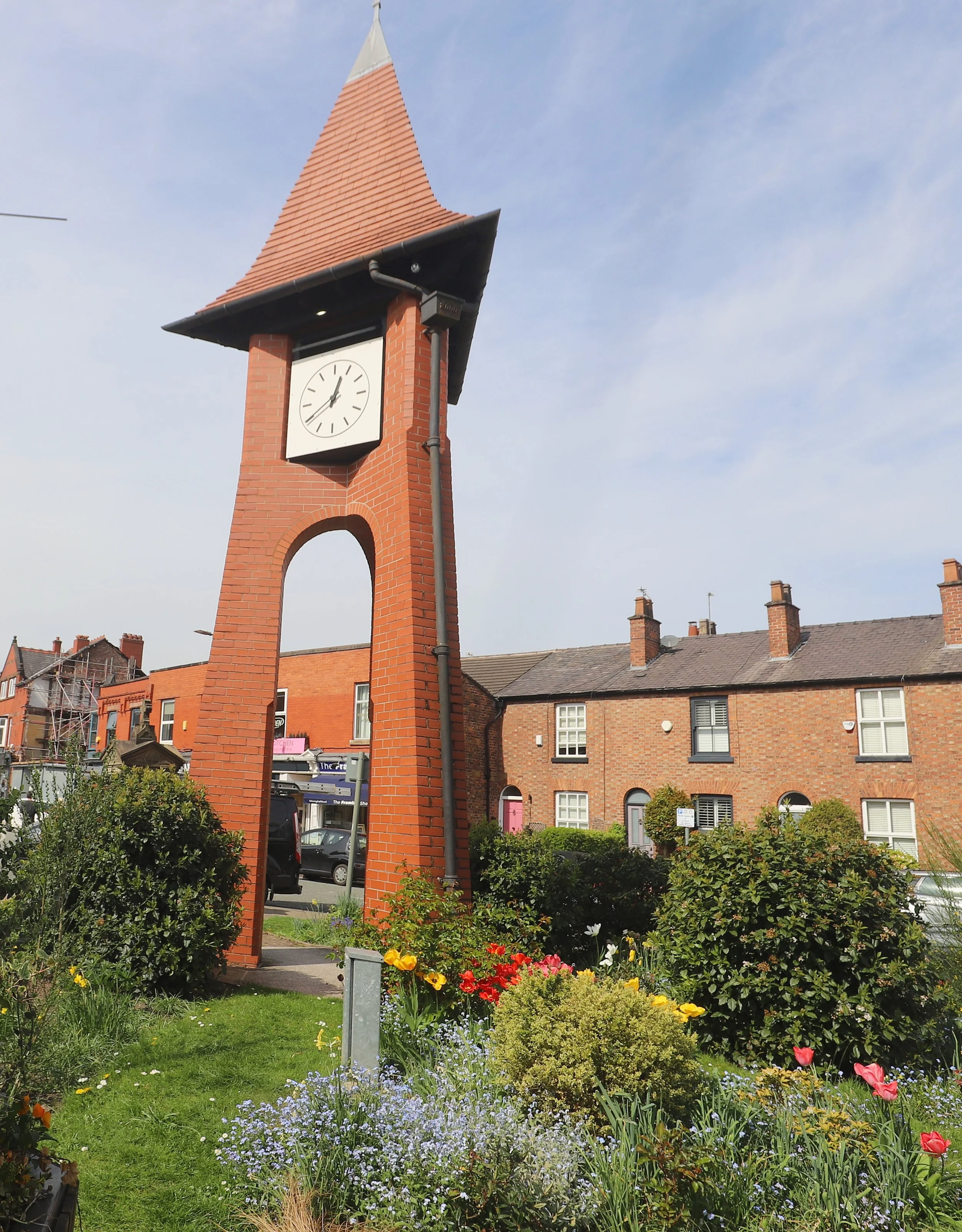 A brick clock tower with a red tiled steep roof and a white clock face showing the time as 5:12, surrounded by a colorful garden with flowers and bushes, and neighboring brick buildings under a partly cloudy sky.