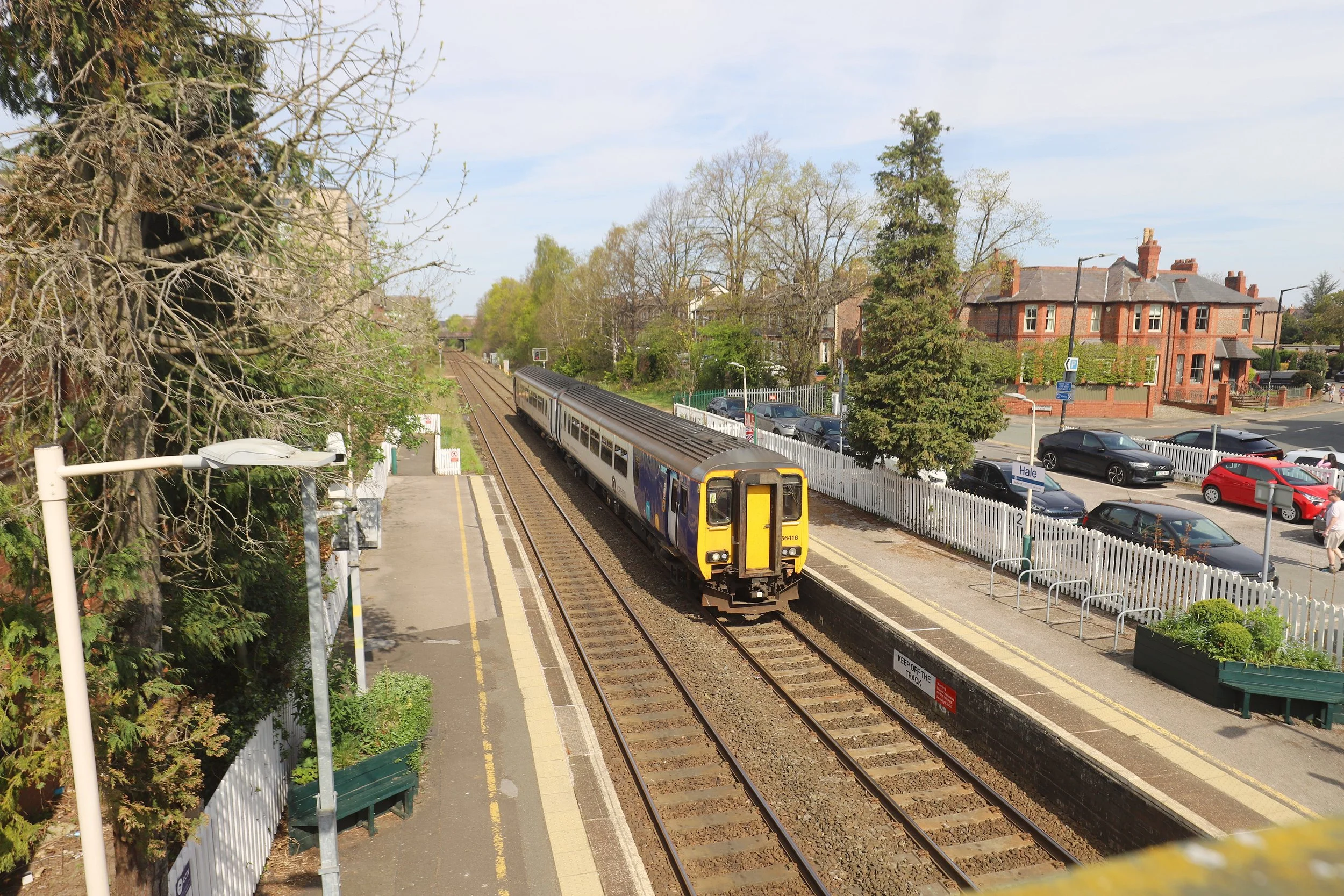 A train at a small train station with a parking area and residential buildings in the background.
