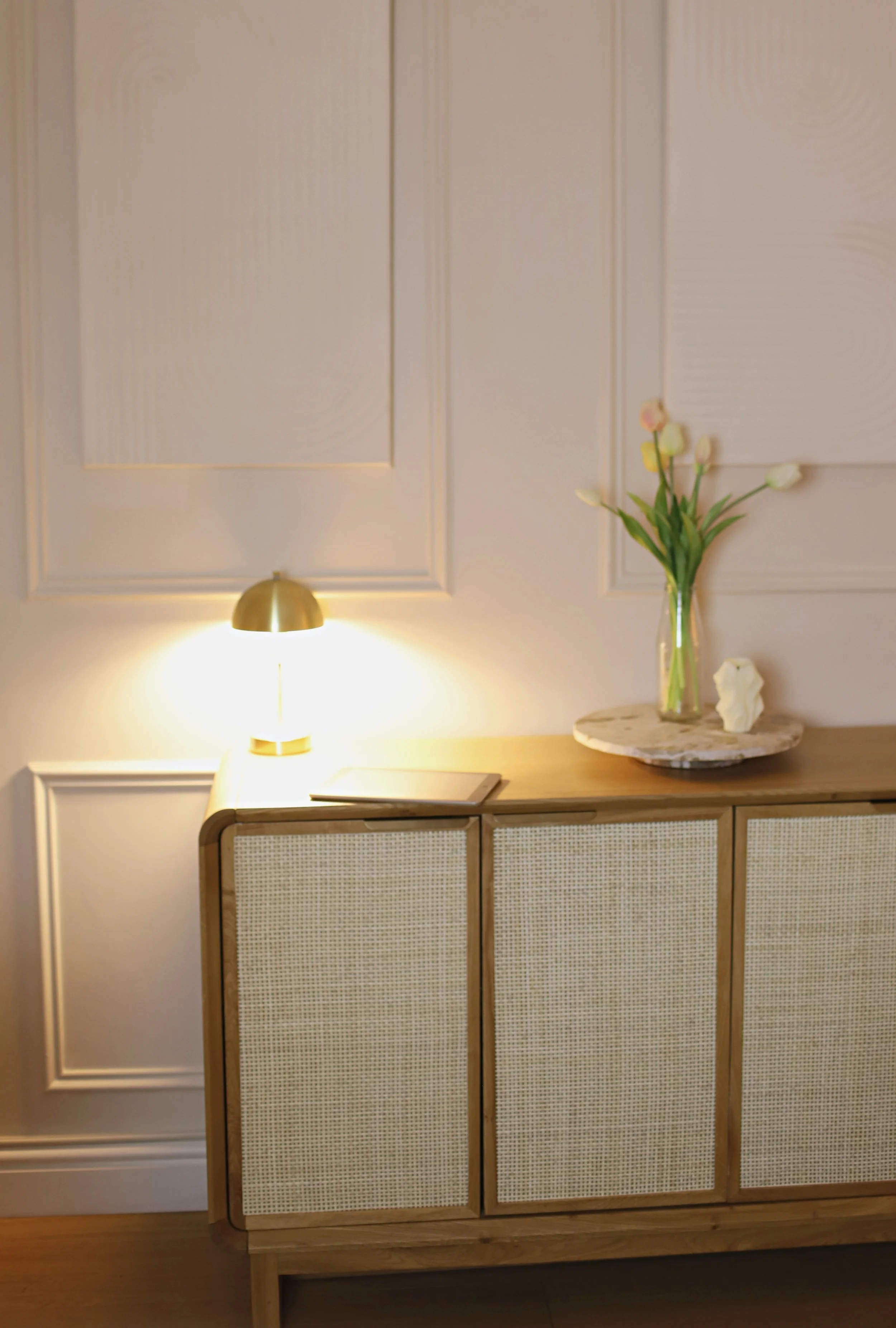 A wooden sideboard with a rattan front, a gold table lamp, a clear glass vase with pink, cream, and white tulips on a marble tray, a white ceramic sculpture, and a book. A wall with decorative molding and beige panels is in the background.