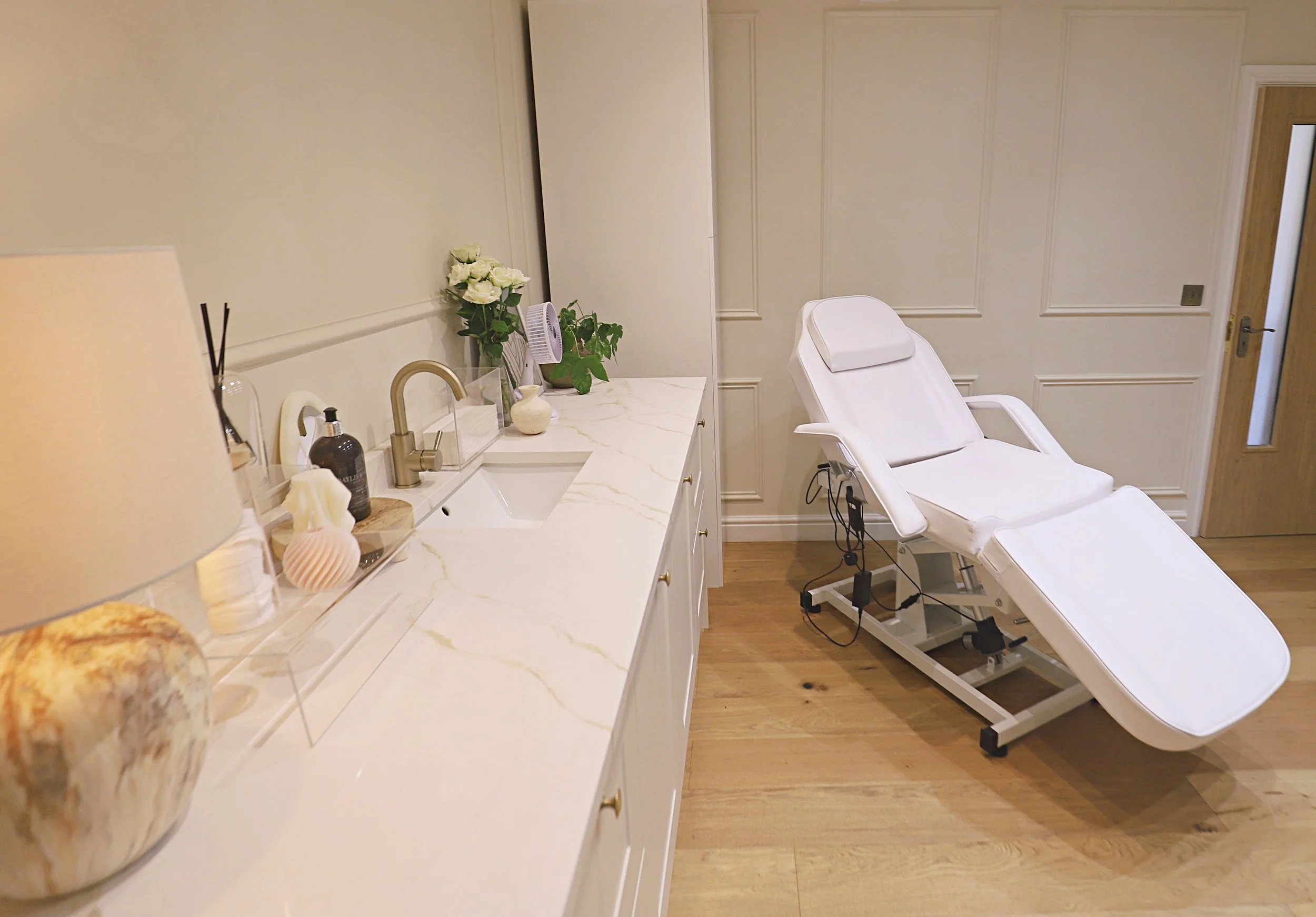 A medical or spa examination room with a white reclining chair, a countertop with decorative items, flowers, and a faucet, and a small fan on the counter.