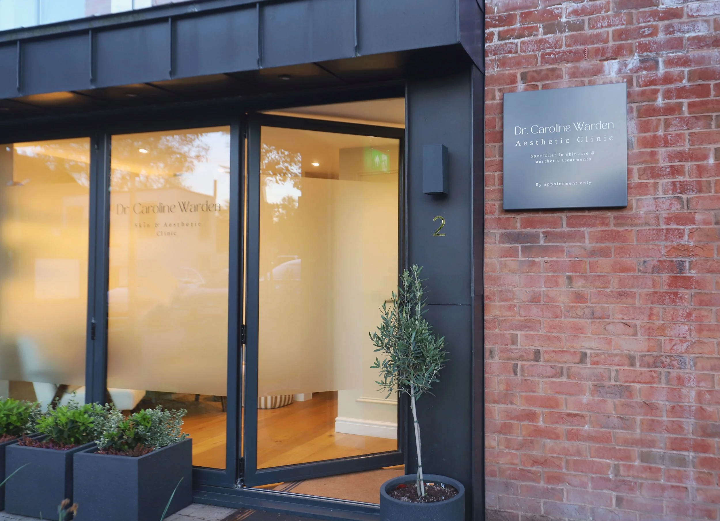 Exterior view of Dr. Caroline Warden Aesthetic Clinic, showing a brick wall with a gray sign indicating the clinic's name, and large glass entrance doors with a reflection of trees and a lit interior.