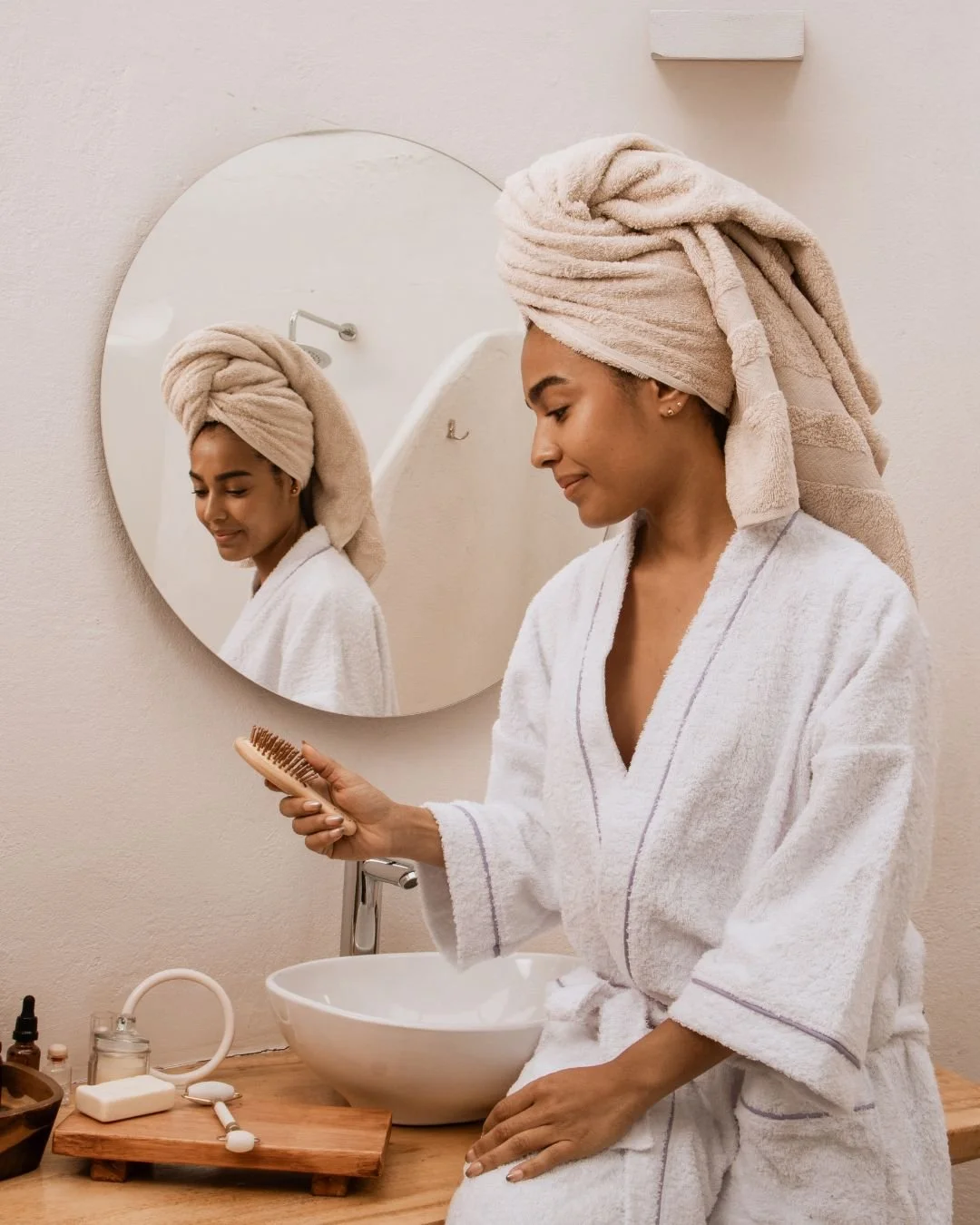 Woman in a towel robe and head wrap brushing her hair in front of a mirror.