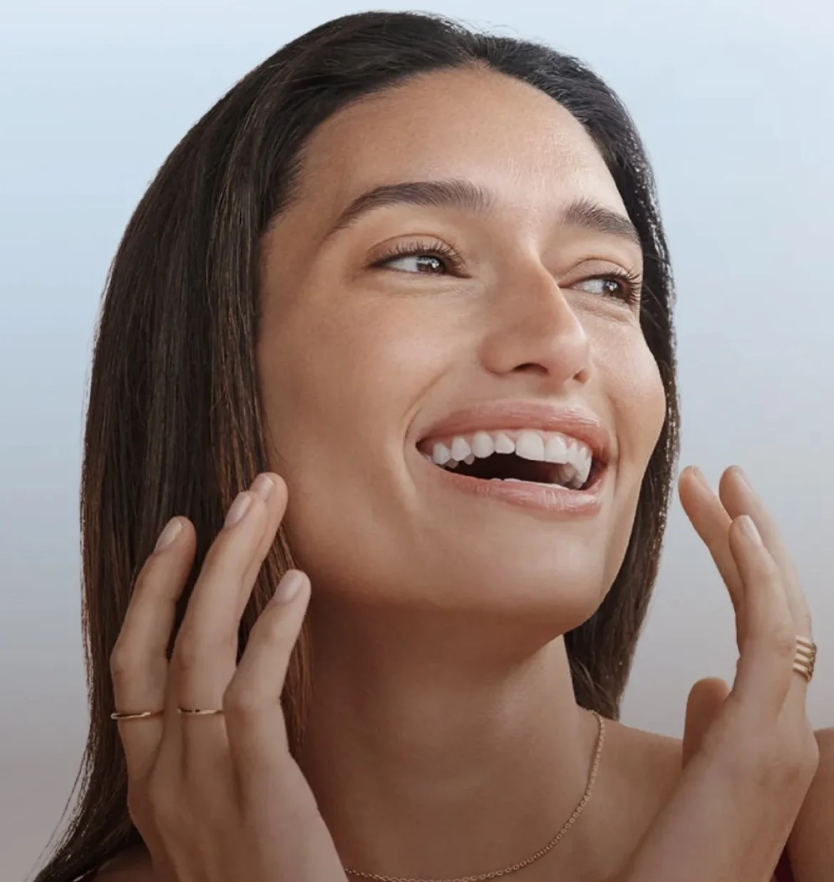 A young woman with long brown hair, smiling and looking up, touching her face with both hands, against a light blue background.