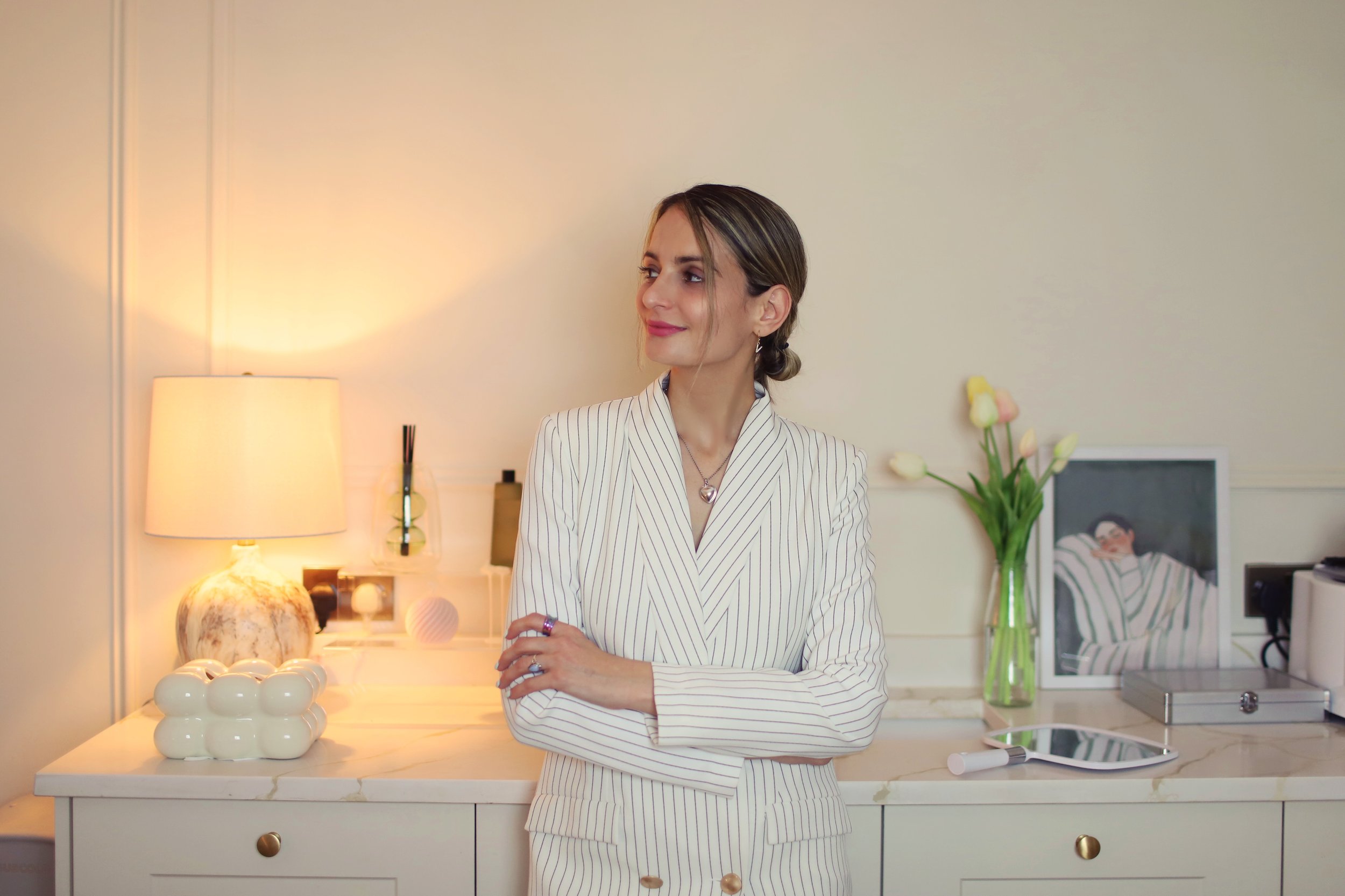A woman with light brown hair styled in a bun, wearing a white and beige striped blazer, stands in a modern kitchen with arms crossed, smiling and looking to her left. The kitchen features a white countertop, a green vase with pink and white tulips, a framed picture on the wall, a beige lamp on the left, and decorative objects including white eggs and a marble lamp base.