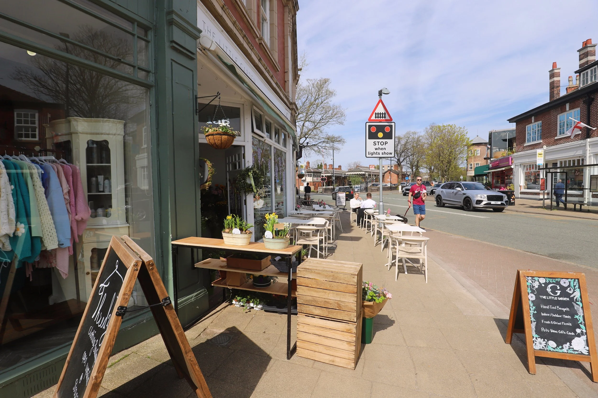 Street scene with outdoor seating area of a café or flower shop, sidewalk, pedestrians, cars, and buildings in the background on a sunny day with blue sky.