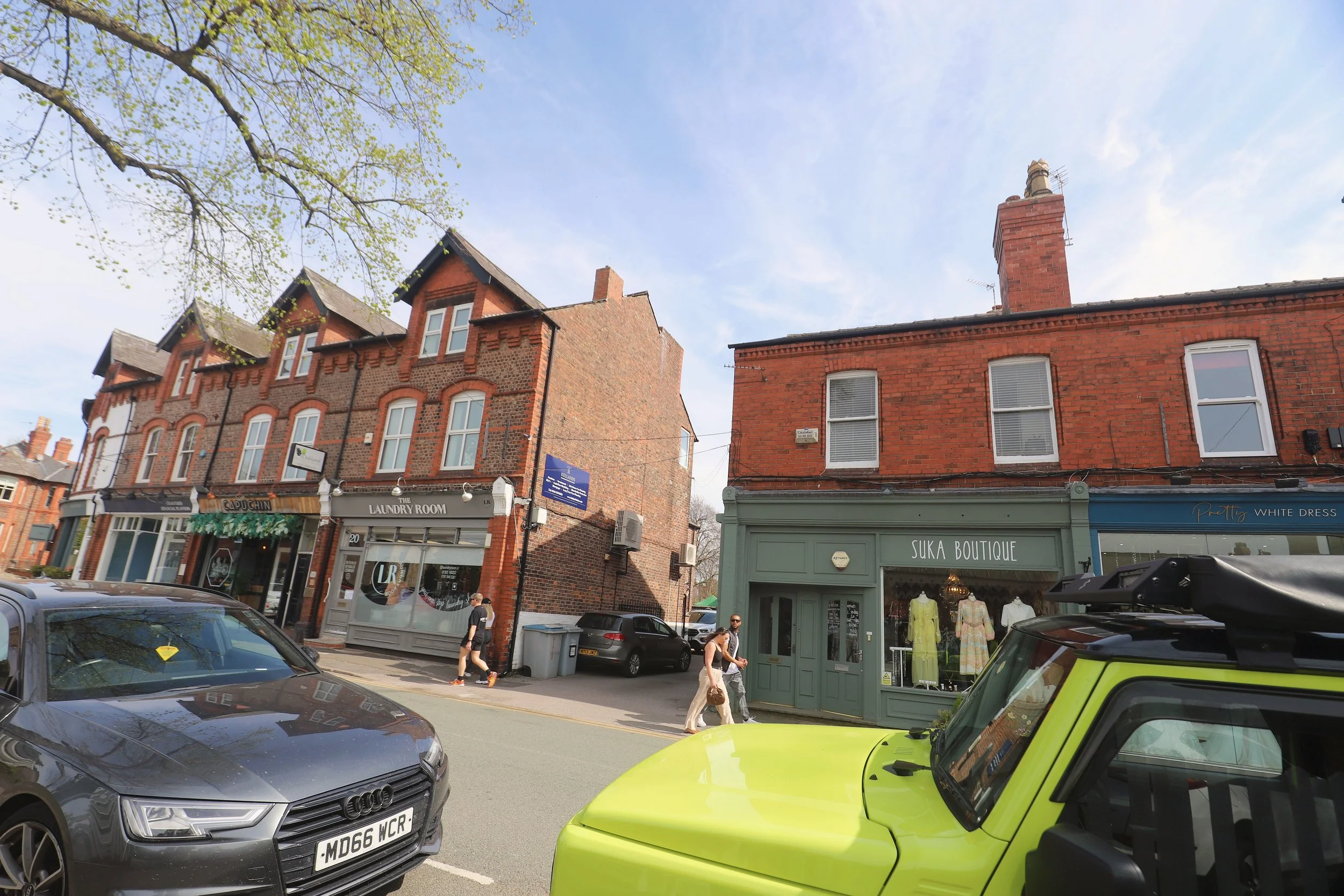 Street view with parked cars, pedestrians walking by, brick buildings with retail shops, trees, and a blue sky with some clouds.