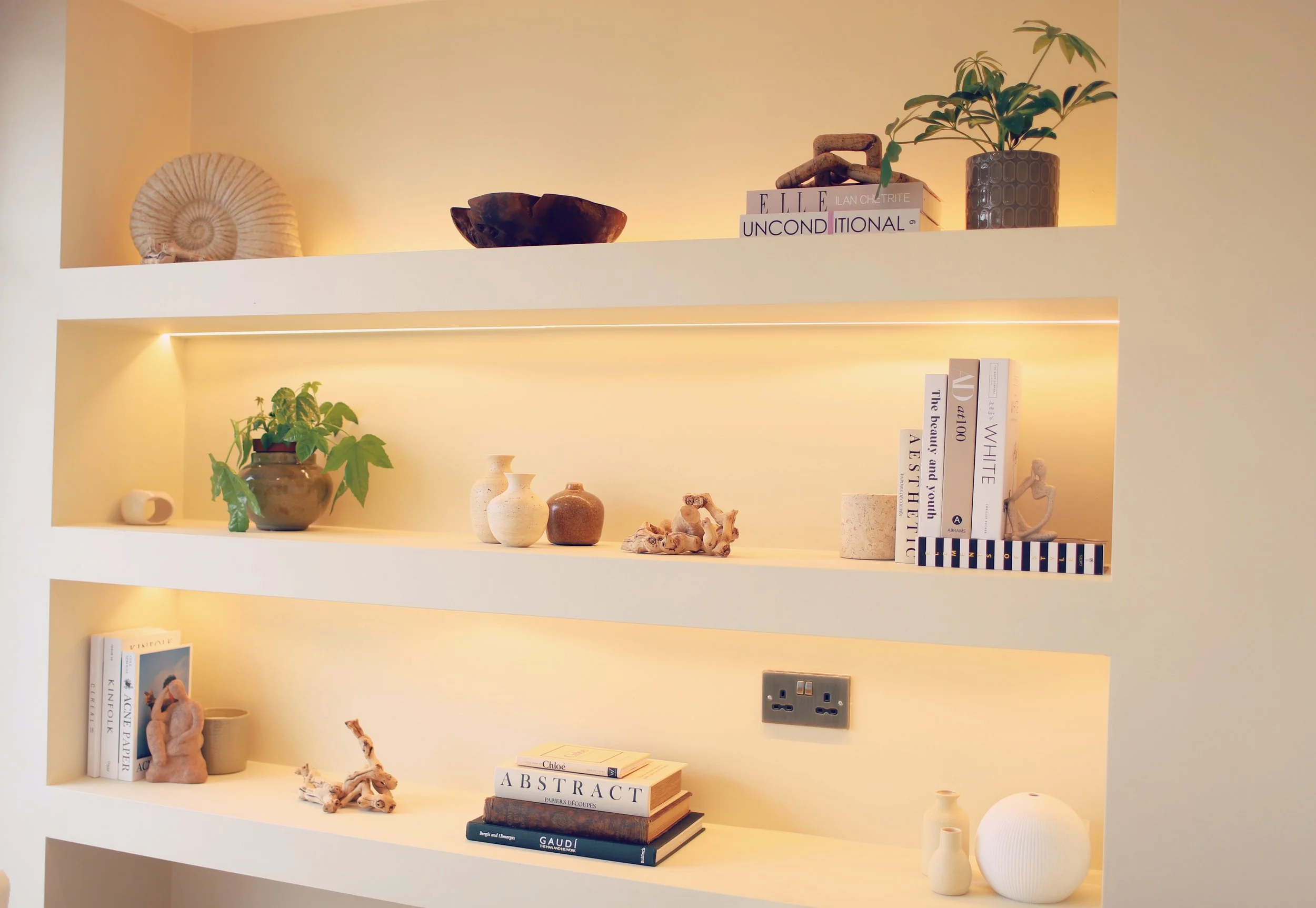 White wall with three built-in shelves, decorated with books, vases, plants, sculptures, and decorative objects, illuminated with warm lighting.