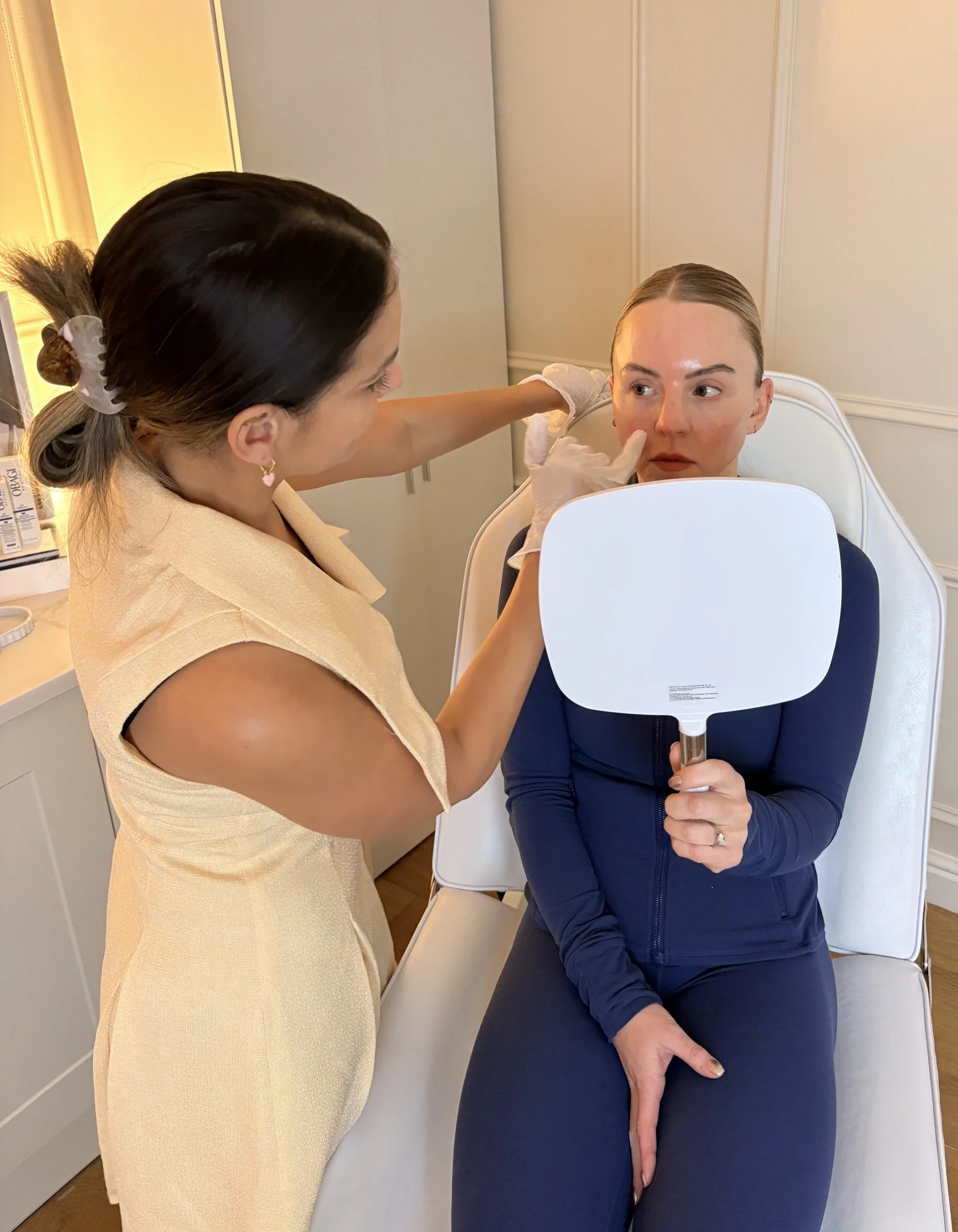 A woman with dark hair tied back, wearing a beige sleeveless top, is examining another woman with light skin and blonde hair, who is sitting in a chair. The sitting woman is wearing a dark blue jacket and is holding a small mirror, while the standing woman points to her face, possibly applying makeup or skincare.