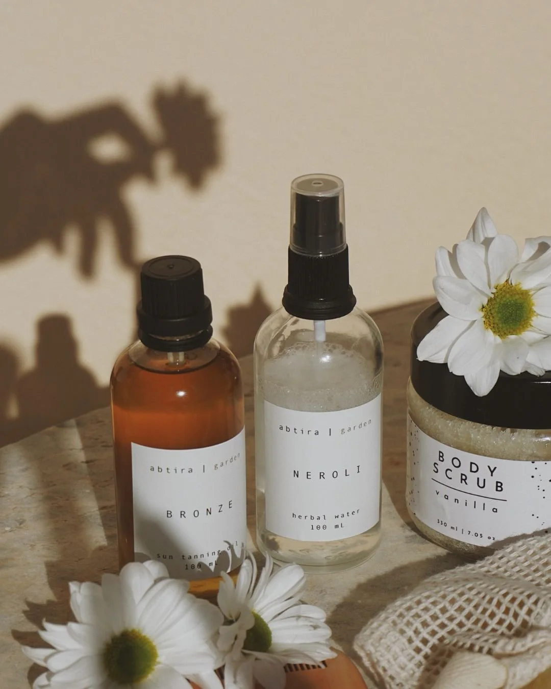 Natural skincare products with body scrub, tanning oil, and herbal water displayed with daisies.