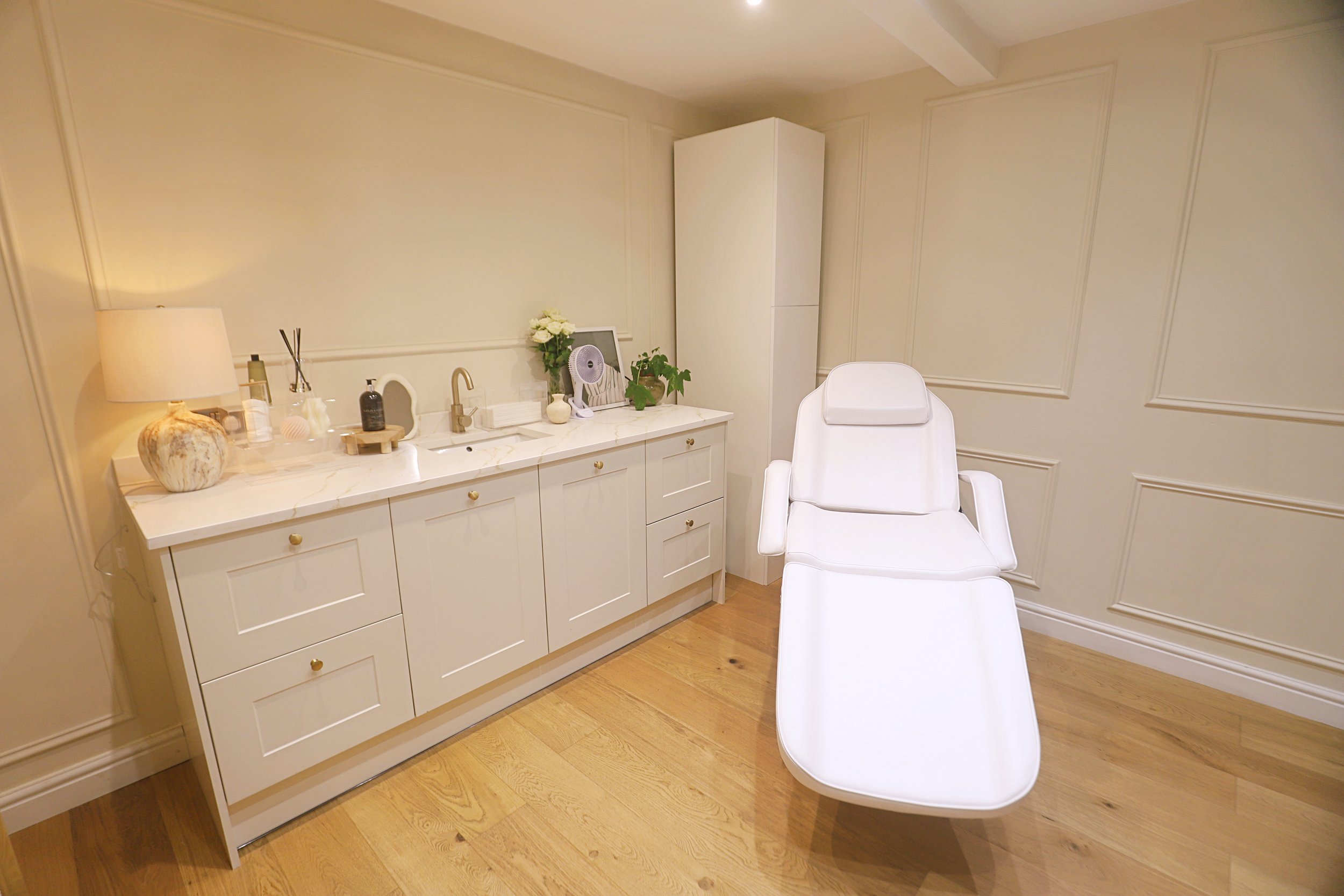 A clinical or spa room with a white reclining chair, a white cabinet with a marble countertop, decorative items, and a vase with flowers.