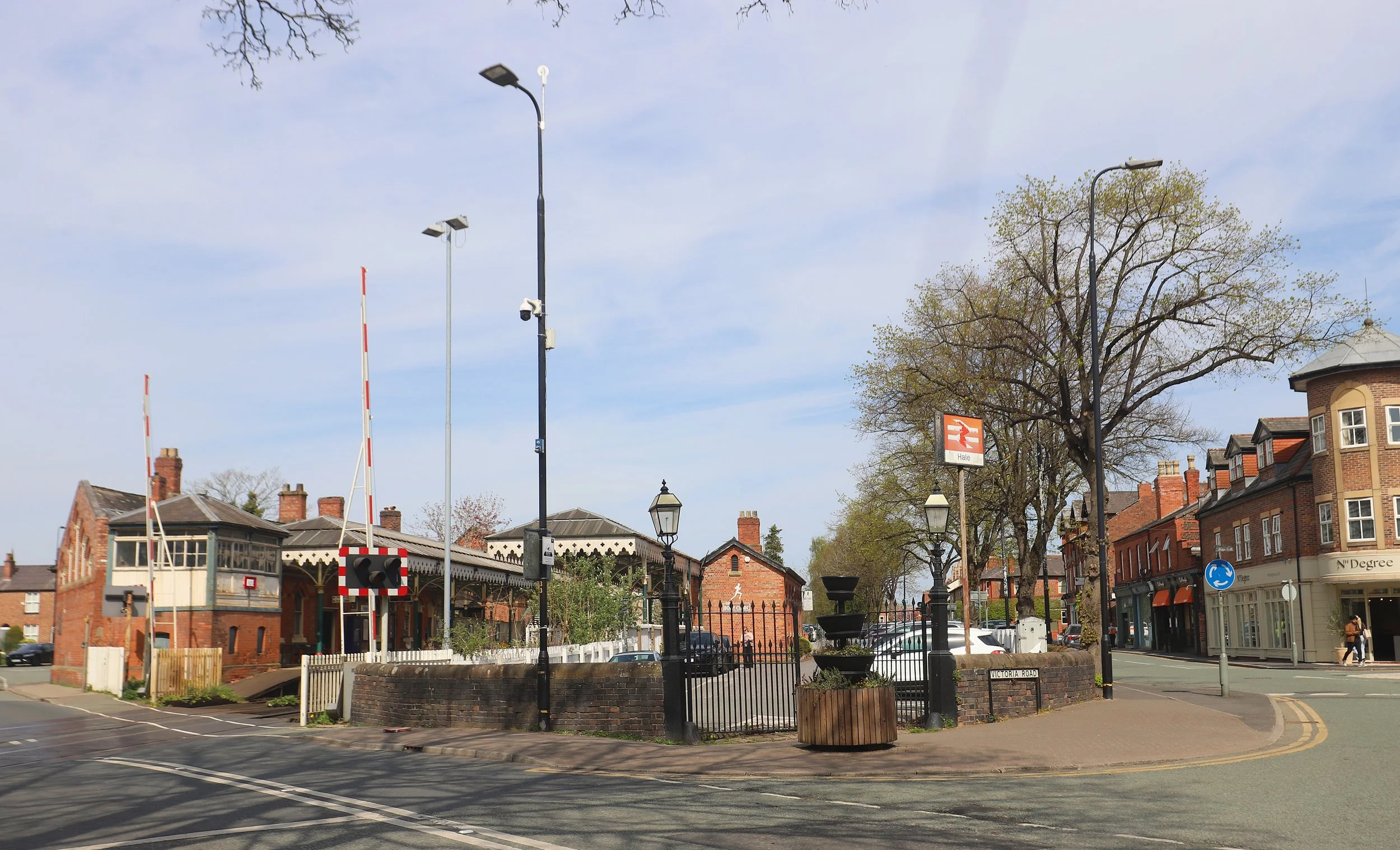 View of a small historic train station with a parking area, trees, and surrounding buildings, under a blue sky with some clouds.