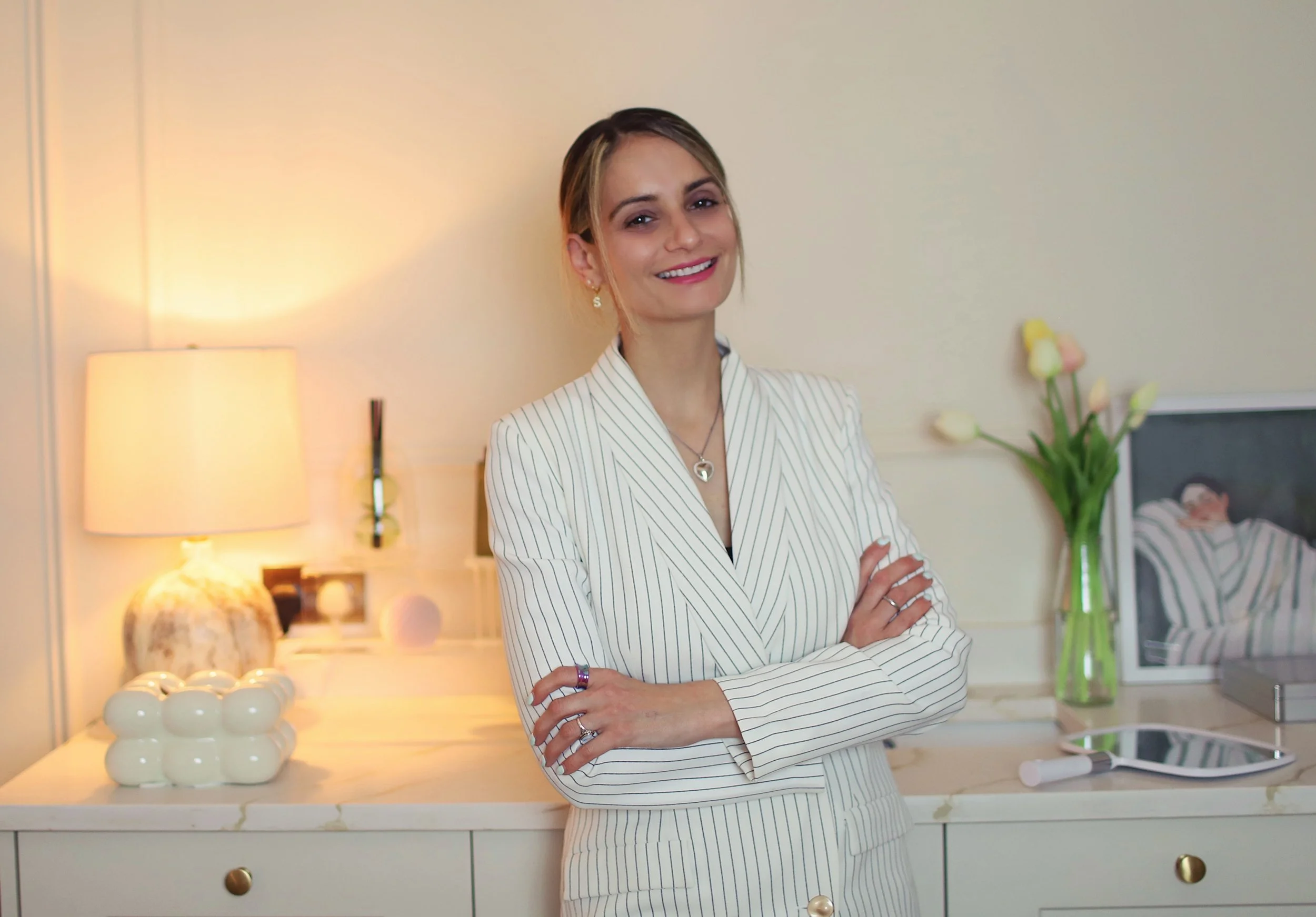 A woman with light skin and blonde hair styled in an updo, wearing a white striped blazer, stands in a warmly lit room with crossed arms, smiling.