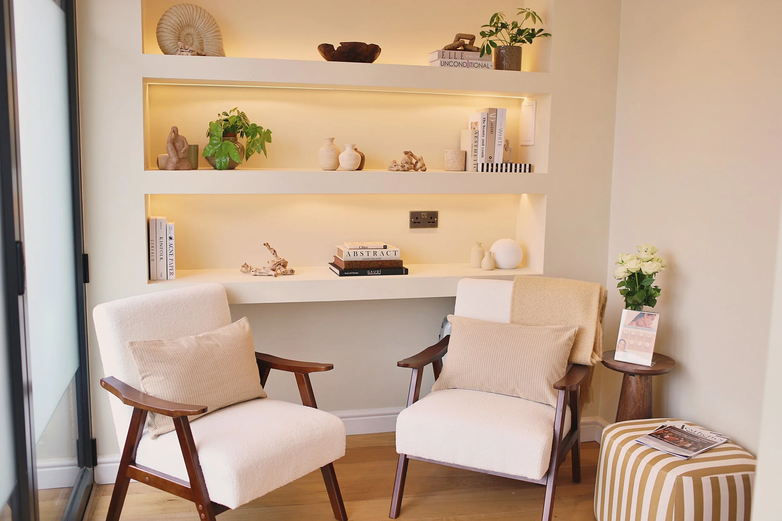 A cozy sitting area with four beige cushioned armchairs with wooden legs and arms, arranged around a small round wooden side table. The background features built-in wall shelves decorated with books, small sculptures, vases, plants, and a spiral shell. There is a window or glass door on the left, and a plant with white flowers on the right.