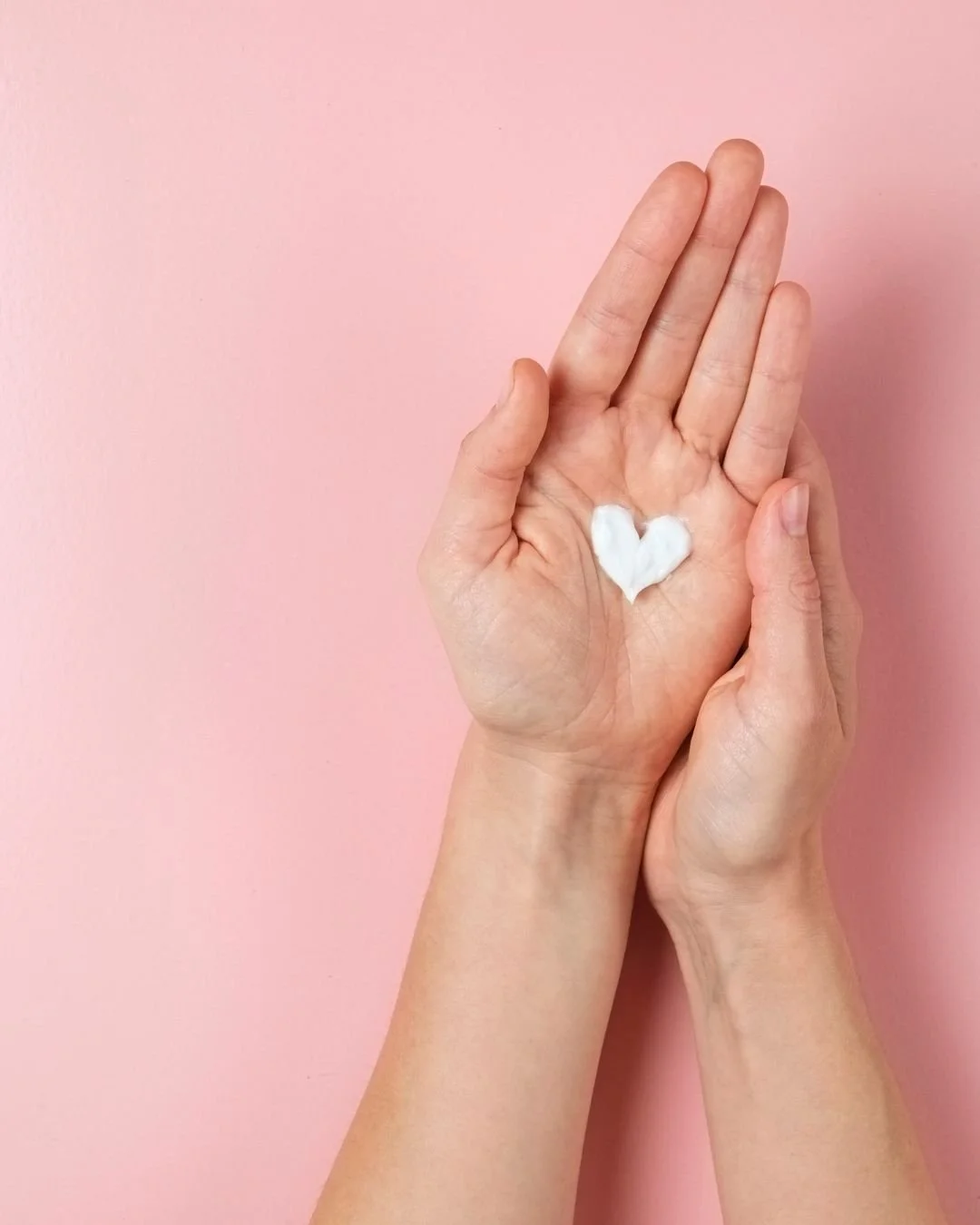 Moisturising cream shaped like a heart on a person’s palm against pink background.