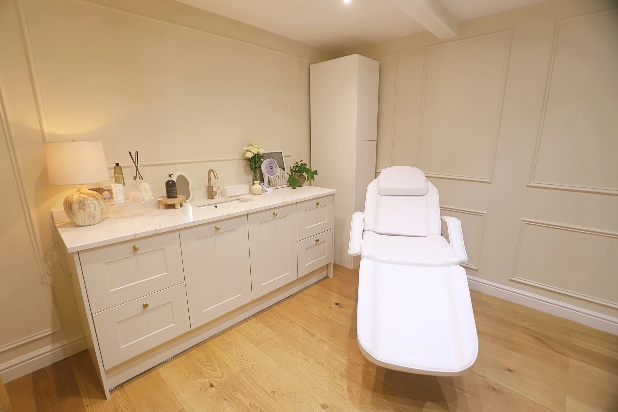 View of a clean and minimal clinical examination room with white cabinetry, decorative items, and a white examination chair.