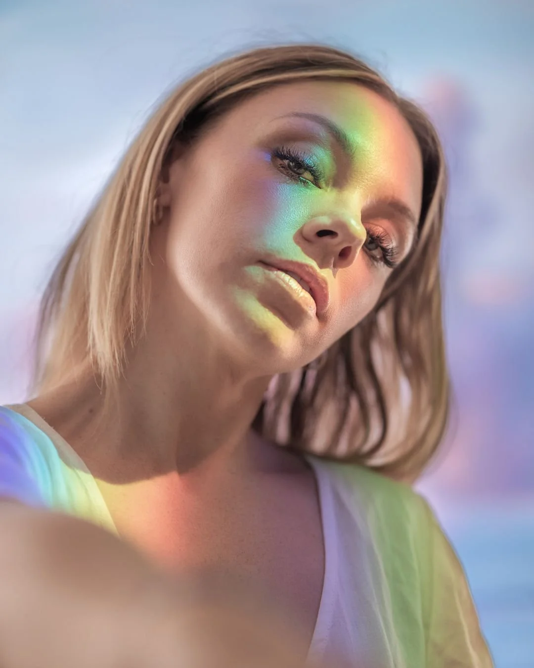 A woman with colorful rainbow-like light projected on her face and shoulder, with her eyes half-closed, looking towards the camera.