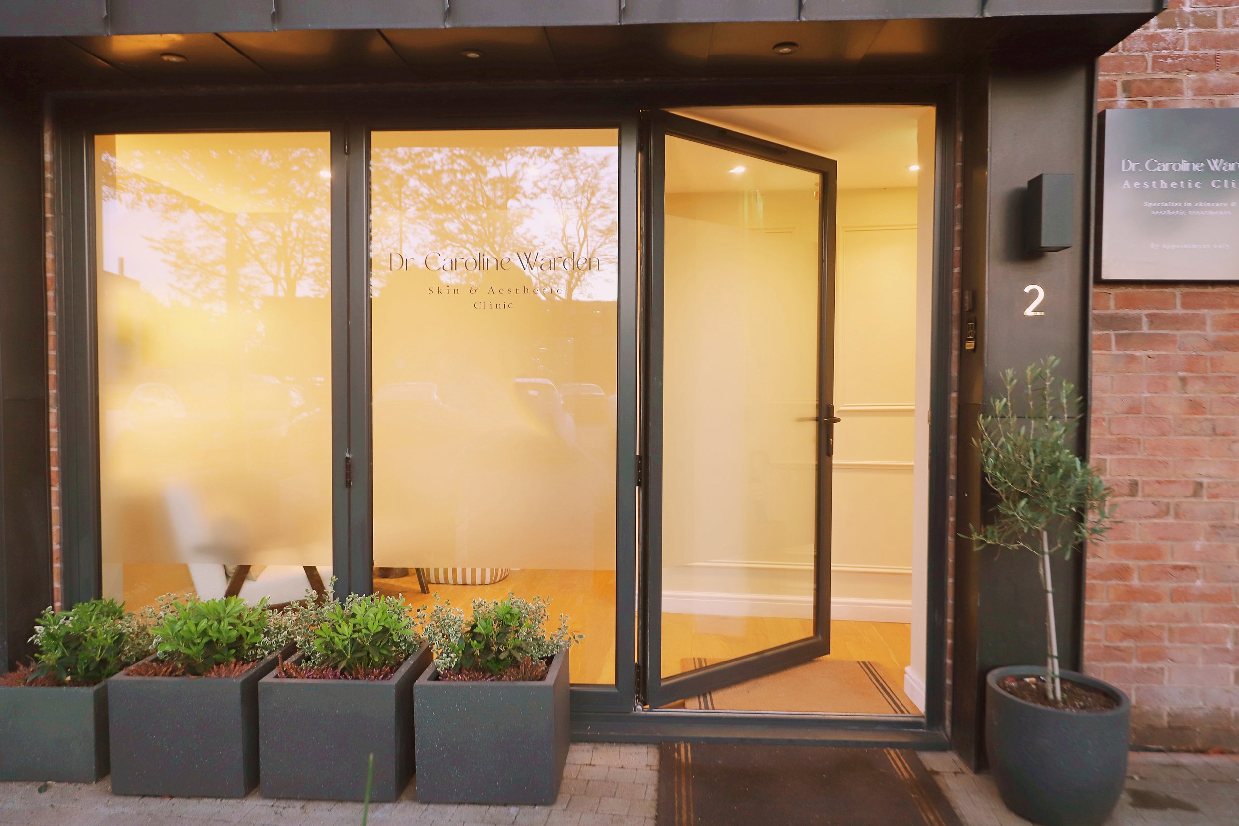 Front entrance of Dr. Caroline Warden Skin & Aesthetic Clinic with glass doors, potted plants, and a black sign on the brick wall.
