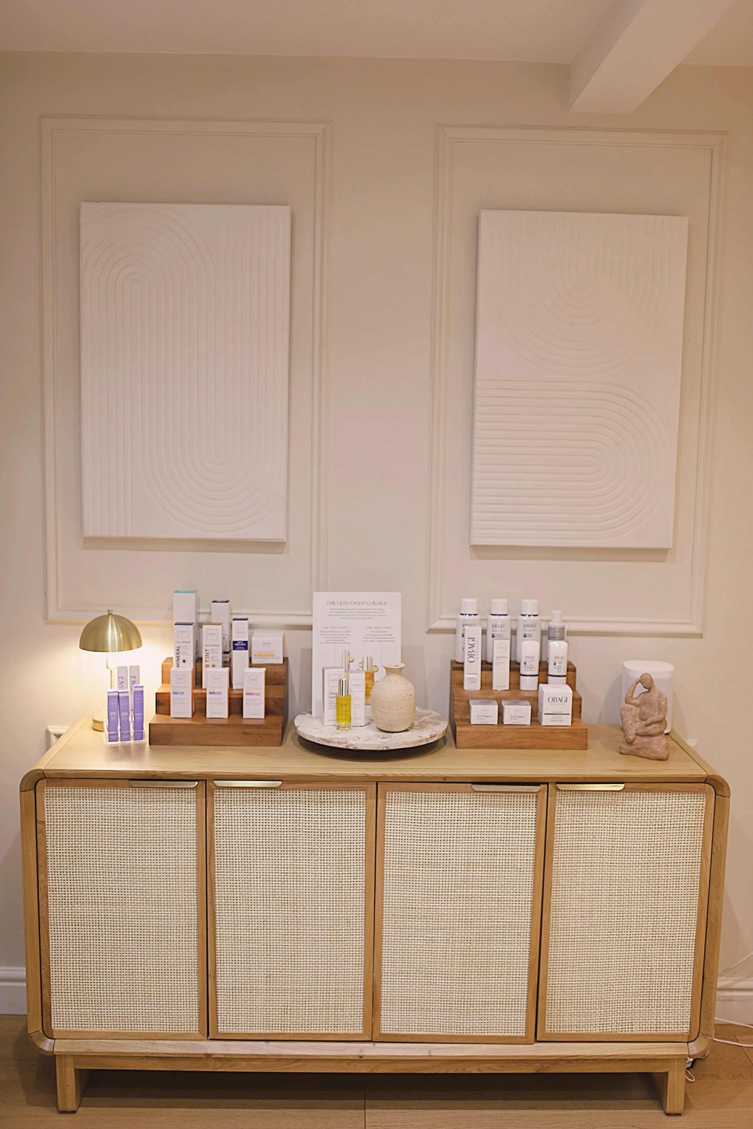 Display of skincare products on a wooden cabinet with two abstract white art pieces on the wall behind. A small lamp with a gold-colored shade on the left and a stone sculpture on the right.