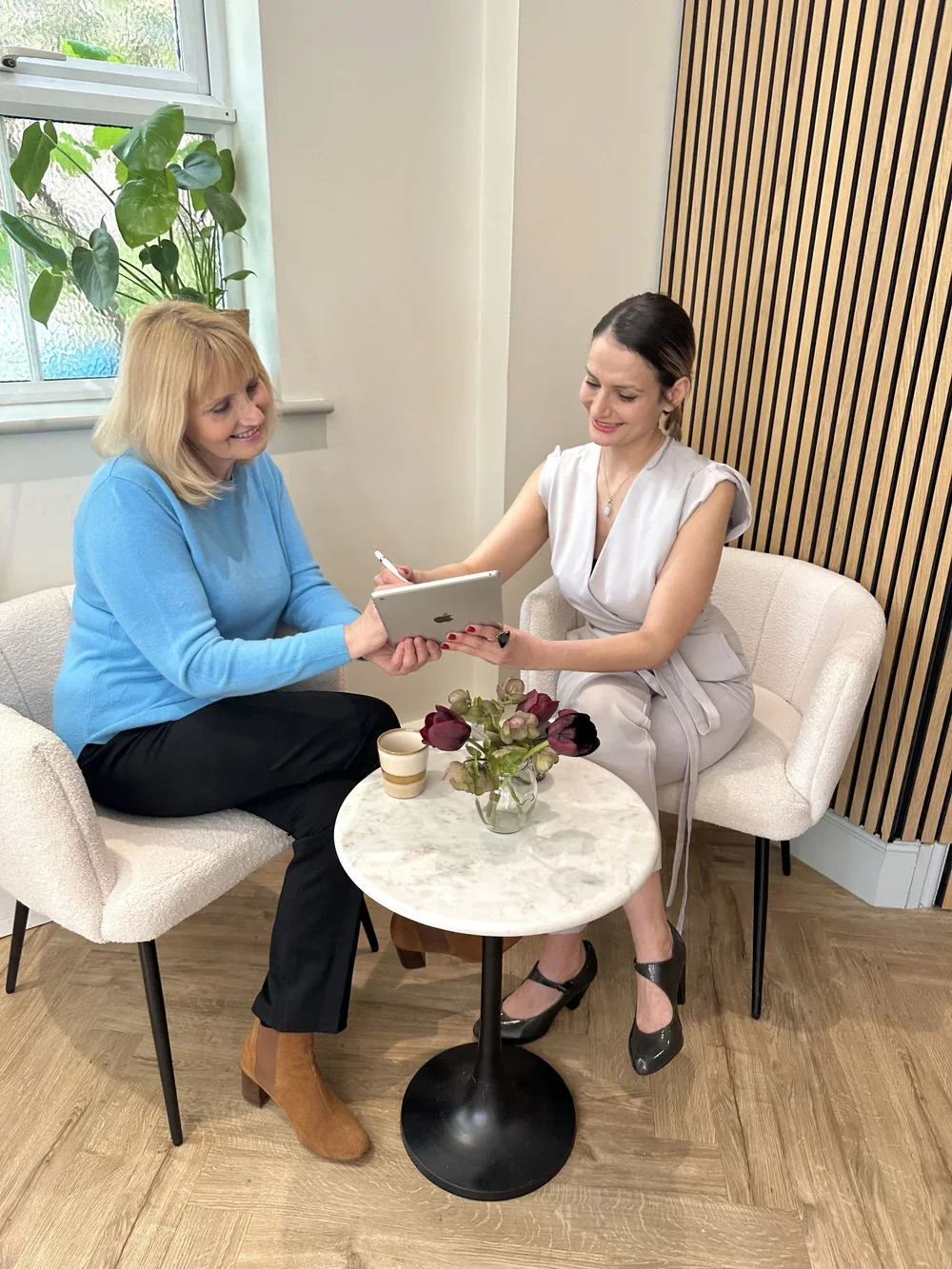 Two women sitting on a couch in a cozy café, sharing a tablet and smiling. One woman with blonde hair wearing a blue sweater, and the other with dark hair in a light gray dress, sitting beside a small round table with a vase of flowers and a cup, near a window with a plant outside.