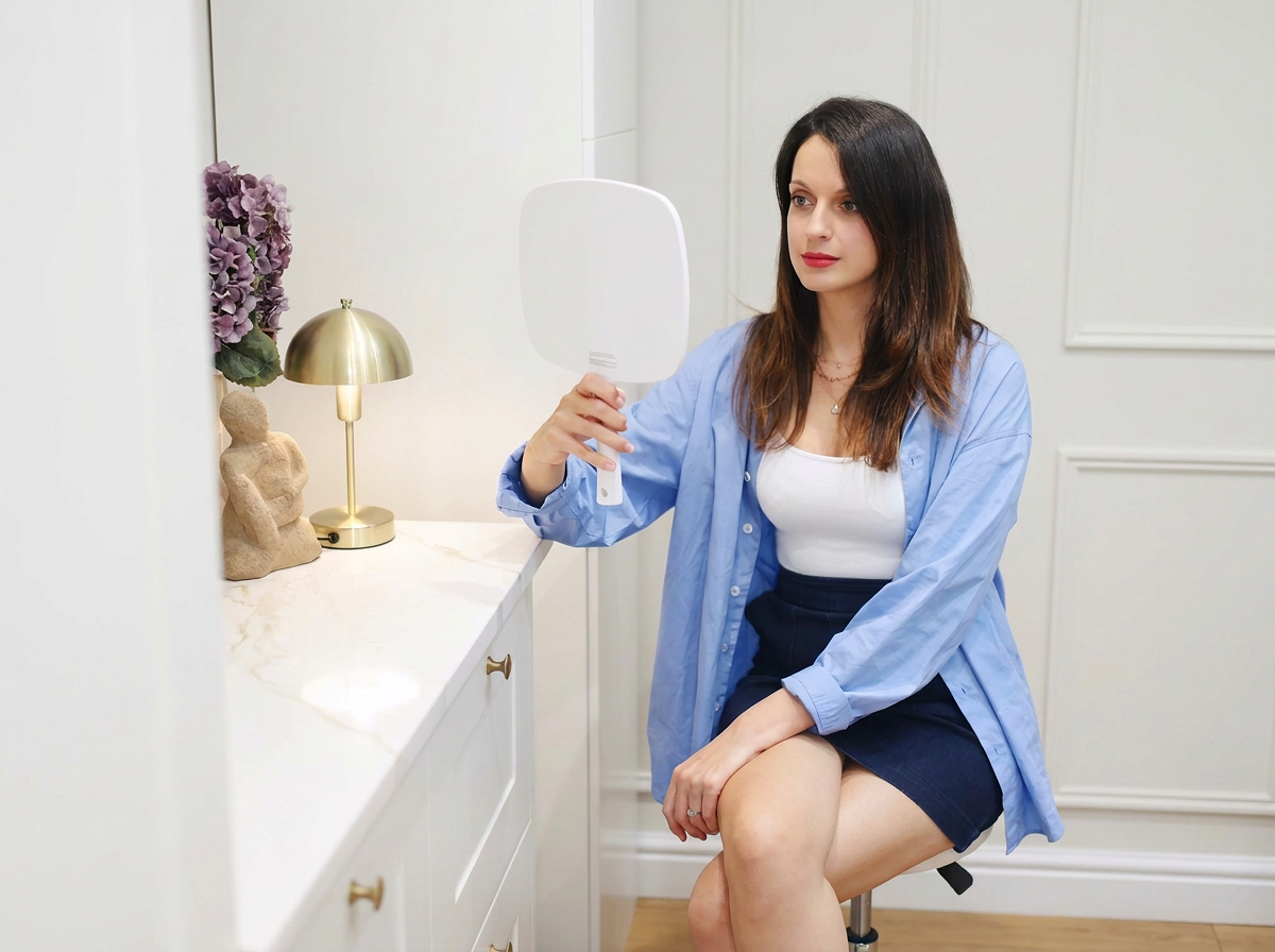 A woman sitting on a chair, holding a hand mirror and looking at her reflection, in a room with white walls and decor such as purple flowers, a gold lamp, and a decorative sculpture.