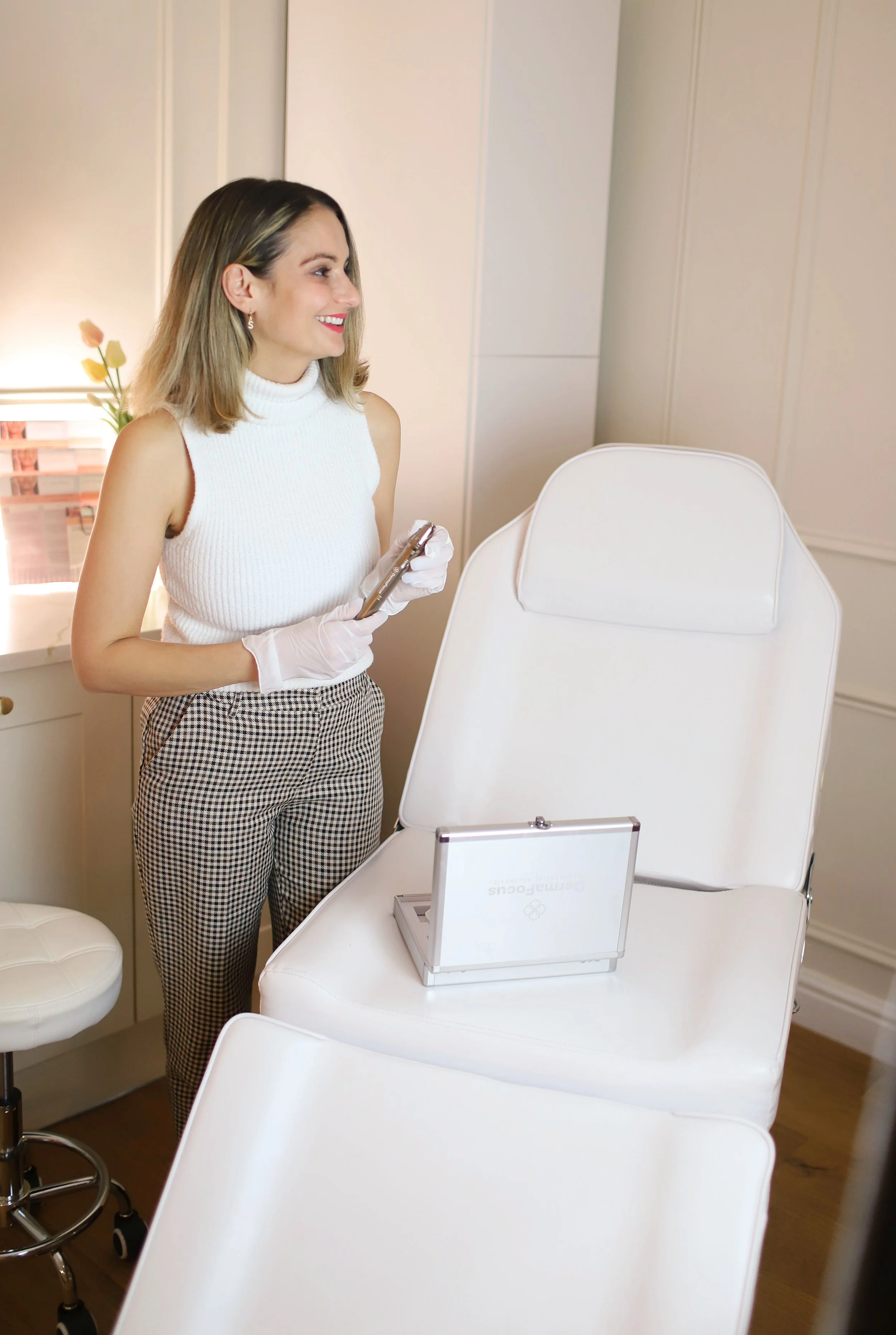 A woman in a sleeveless white top and checkered pants is smiling and holding a phone, standing next to a white beauty chair with a mirror on it, in a room with soft lighting and white walls.