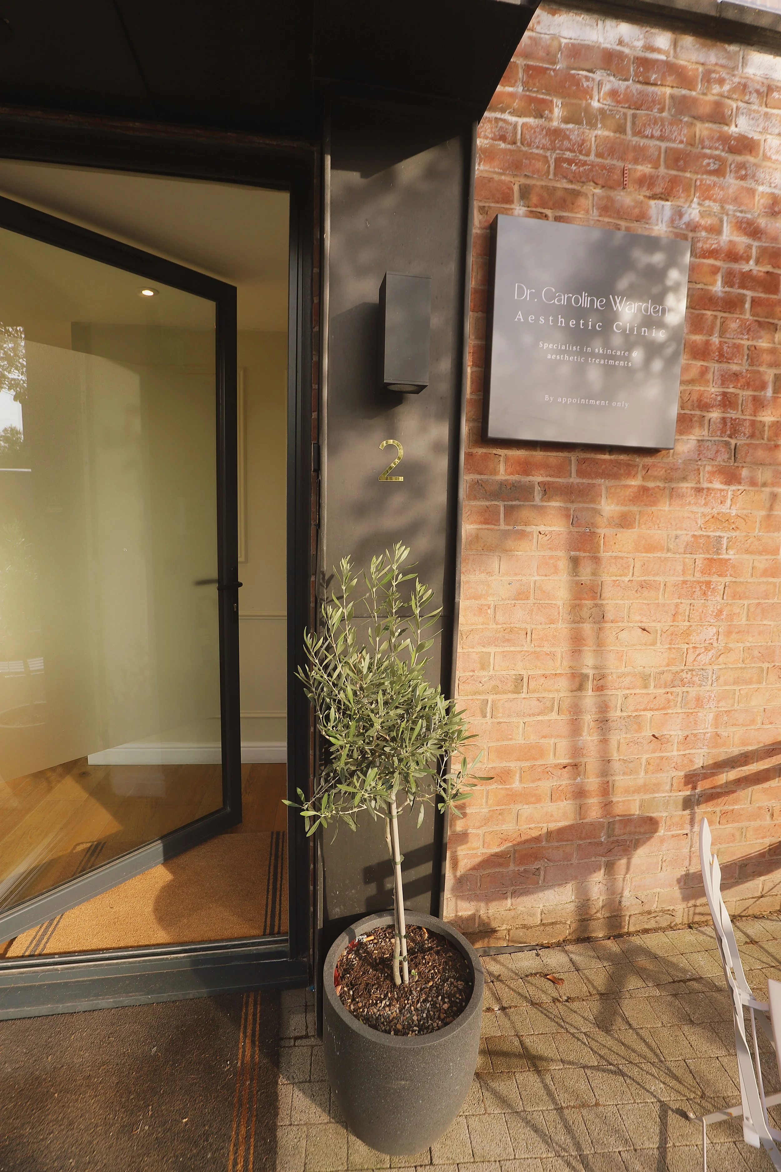 A potted olive tree in front of a brick wall next to a glass door with a black frame at the entrance of a clinic called Dr. Caroline Warden Aesthetic Clinic, which offers skincare and aesthetic treatments, by appointment only.