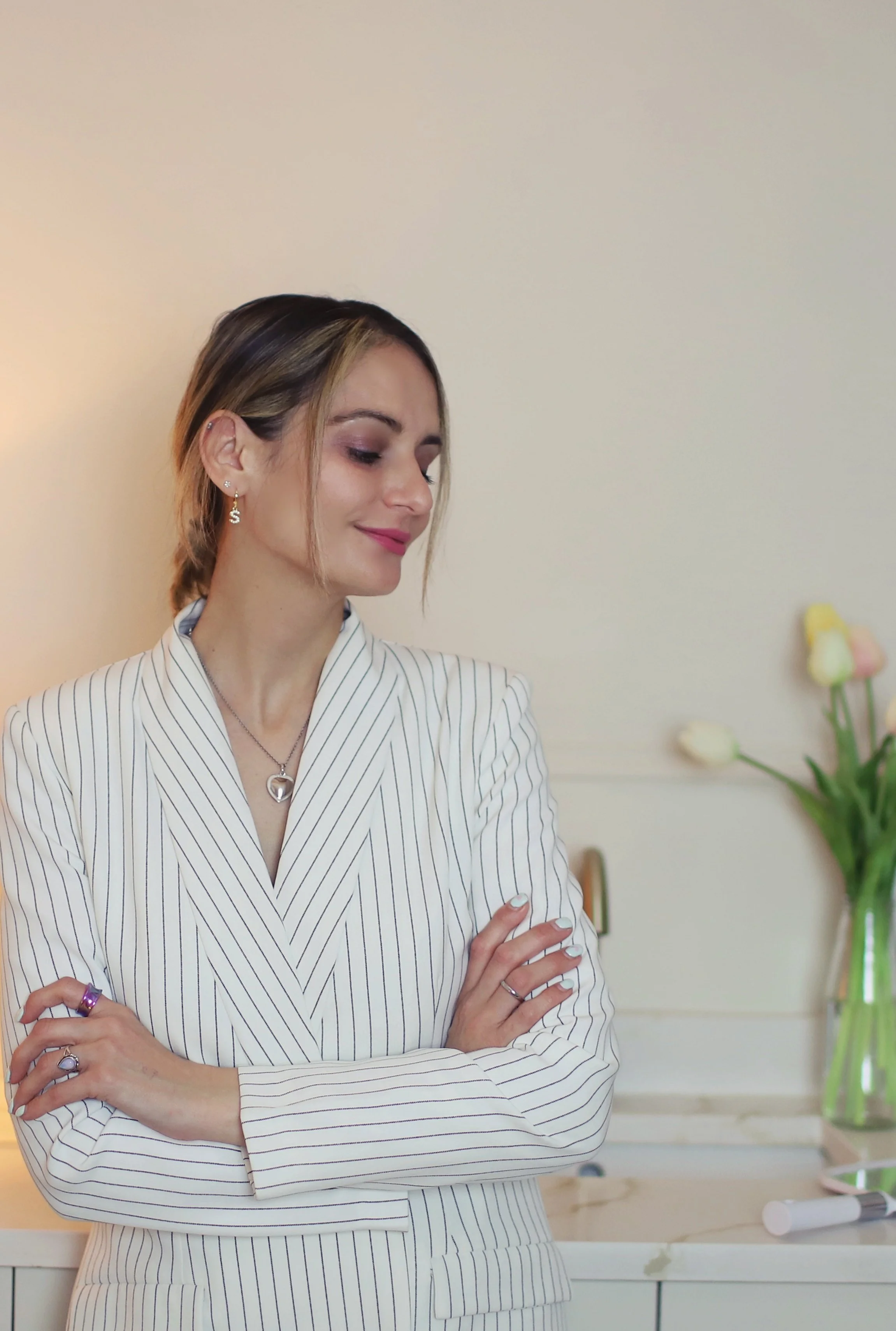 A woman with short brown hair, wearing a white pinstripe blazer, is smiling with her eyes closed, arms crossed, and standing indoors near a vase of tulips.