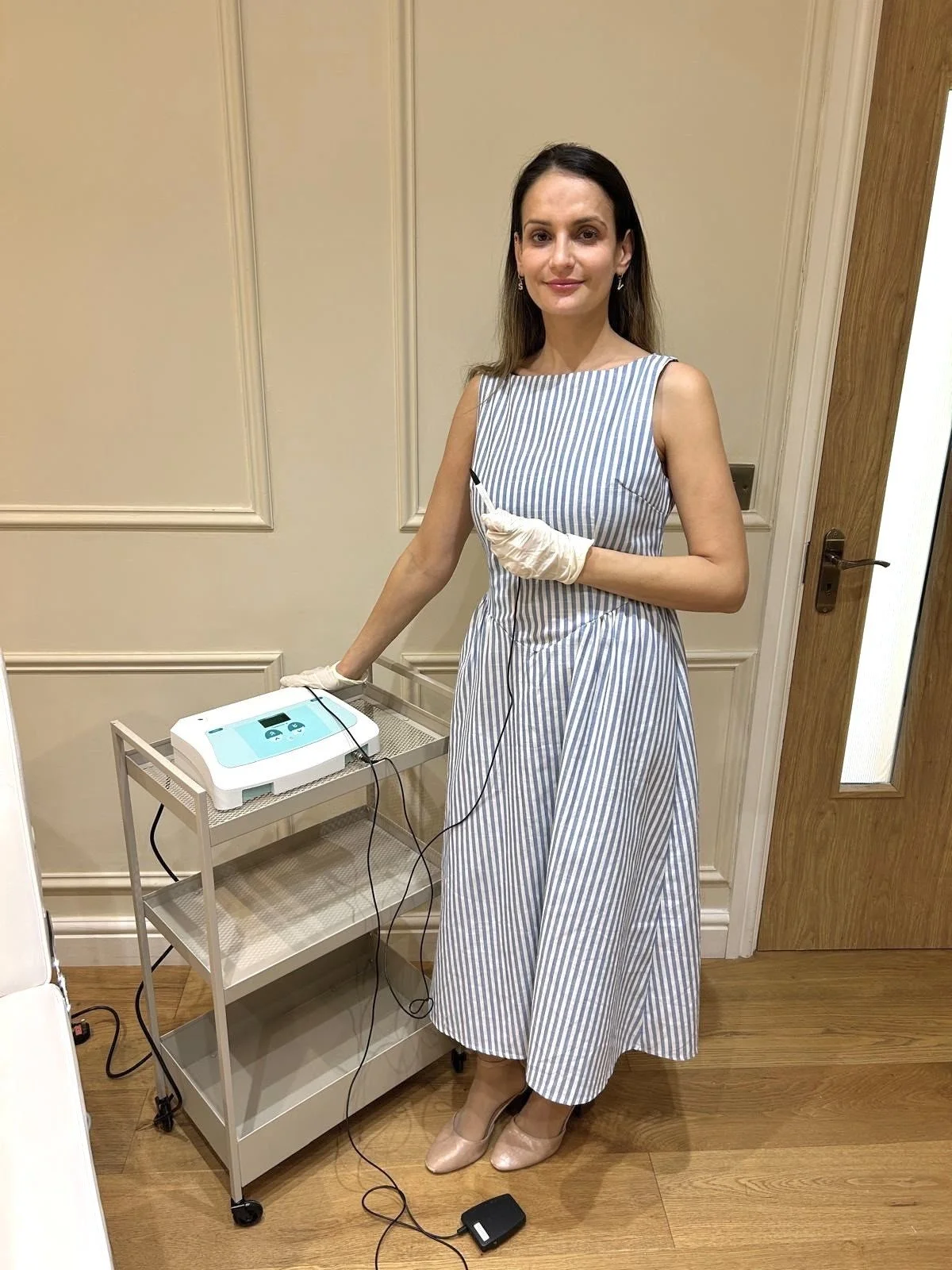 A woman in a striped dress standing next to medical equipment, wearing gloves and holding a cable, in a clinical setting.