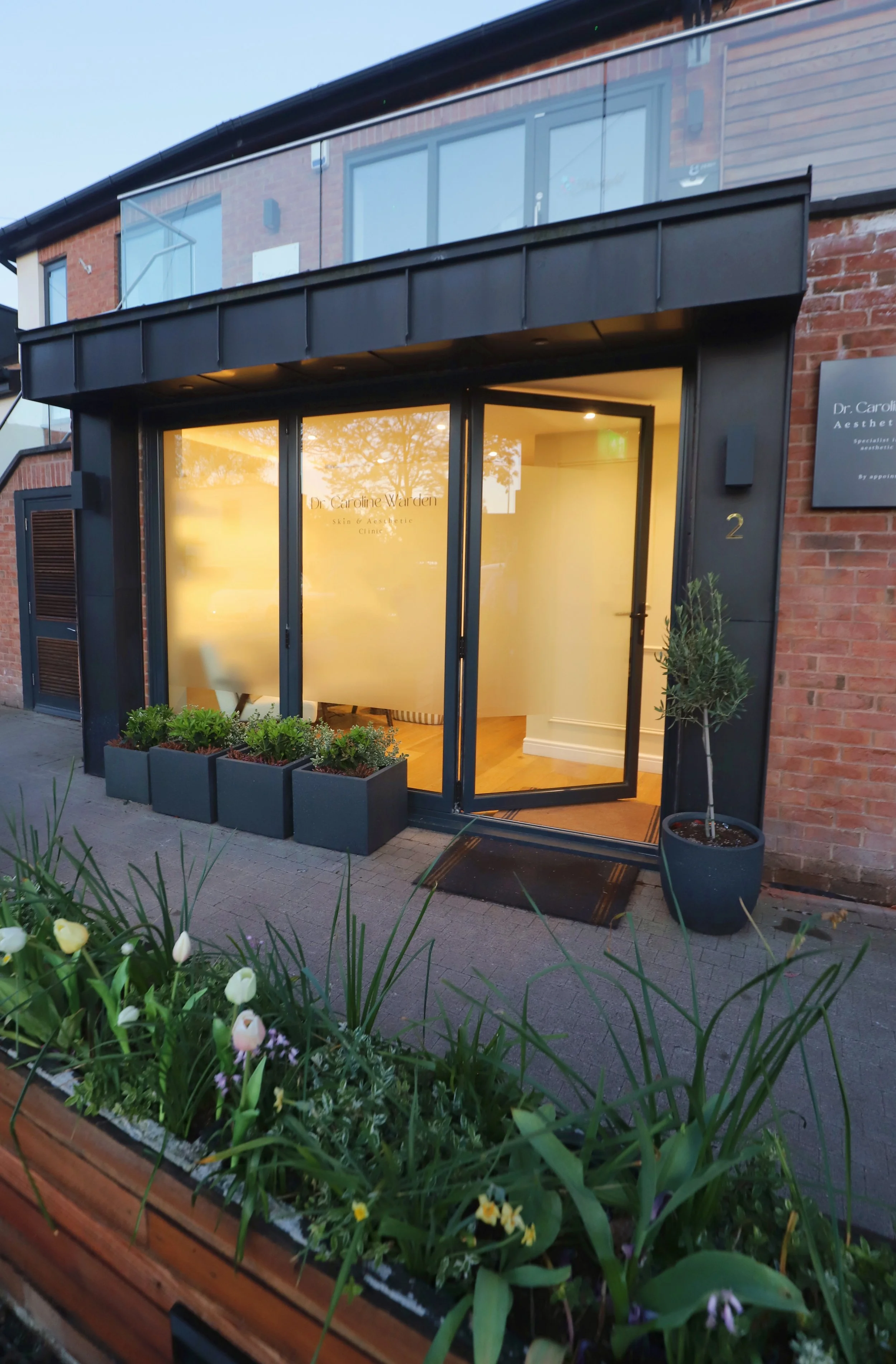 The exterior of a wellness clinic with glass doors and window, potted plants outside, and a flower bed with tulips and other flowers.