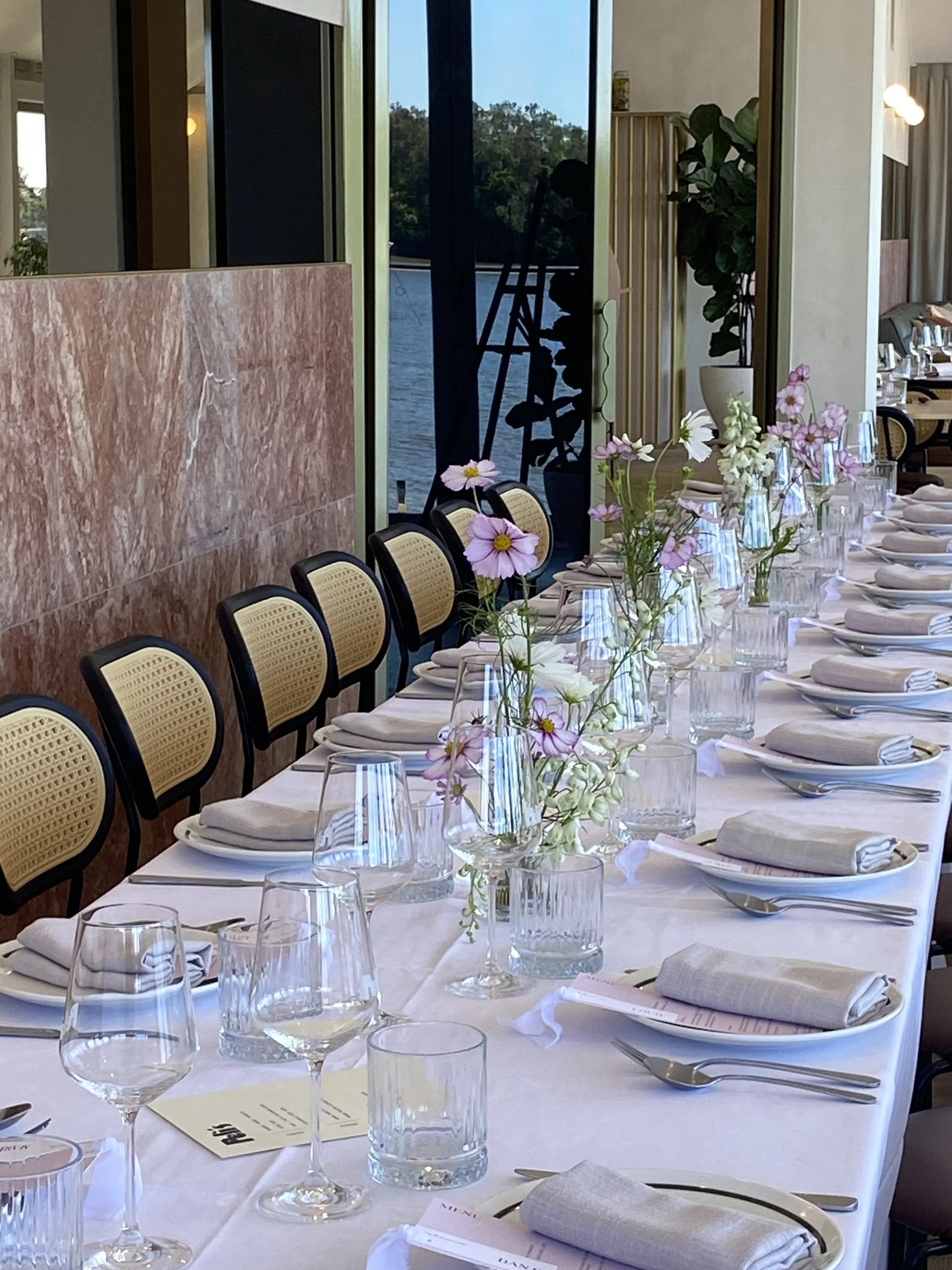 A long dining table set for a formal meal with white tablecloths, stacked plates, neatly folded napkins, wine glasses, and vases with pink and white flowers. In the background, large windows reveal a view of water and trees outside.