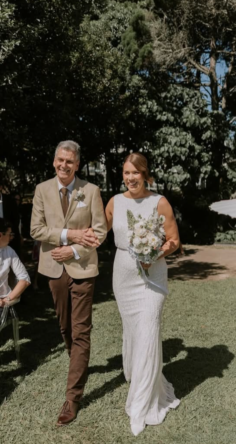 A bride in a white lace wedding dress holding a bouquet walks outside with an older man in a tan suit and brown pants, both smiling. They are under large trees on a sunny day.