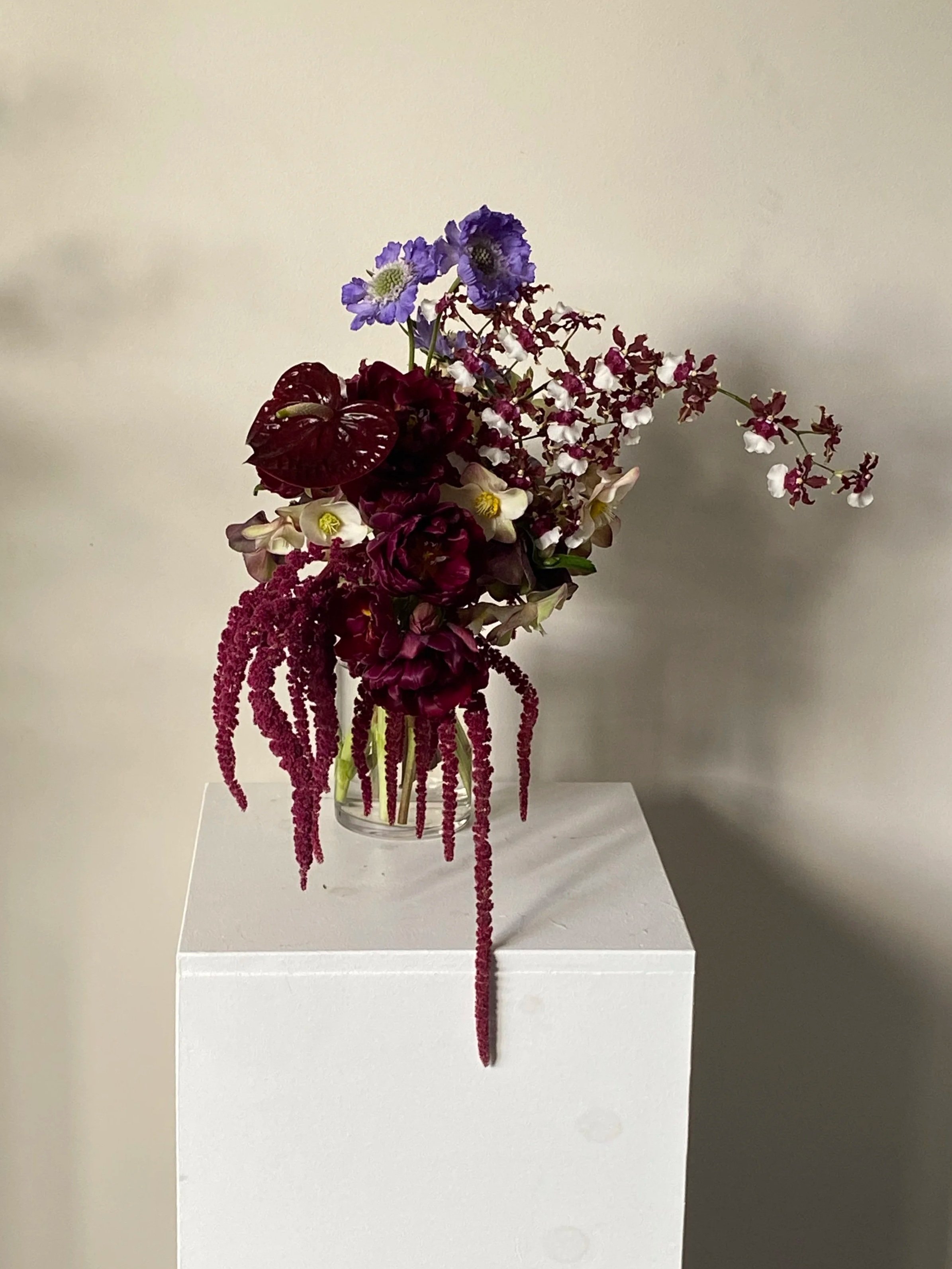 A floral arrangement in a clear glass vase on a white pedestal against a plain wall, featuring purple, white, and deep red flowers with trailing red amaranth.