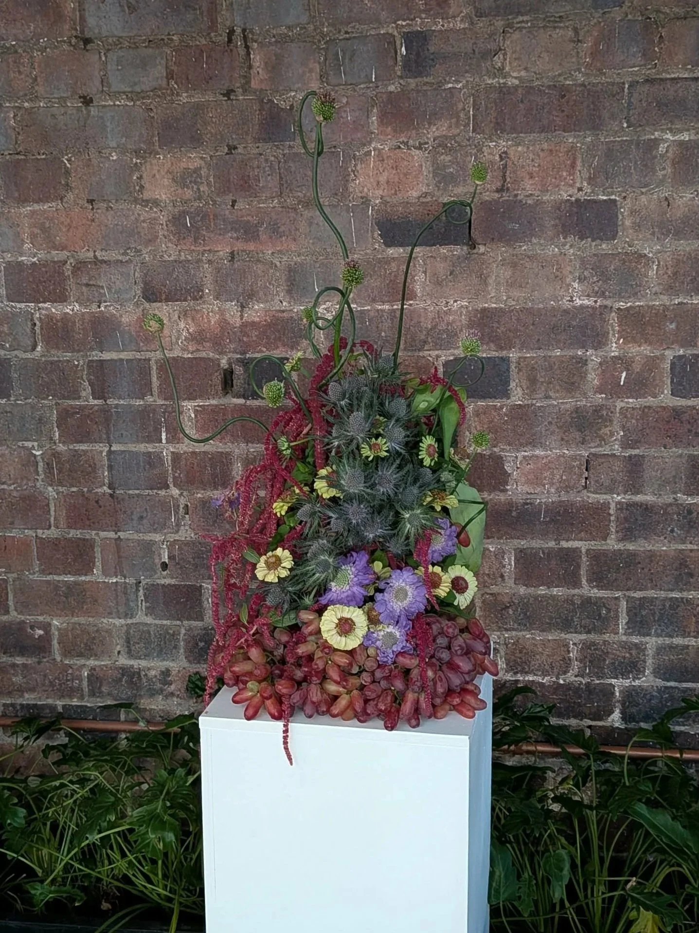 A vibrant floral arrangement featuring various colorful flowers and green foliage, displayed against a brick wall backdrop on a white pedestal.
