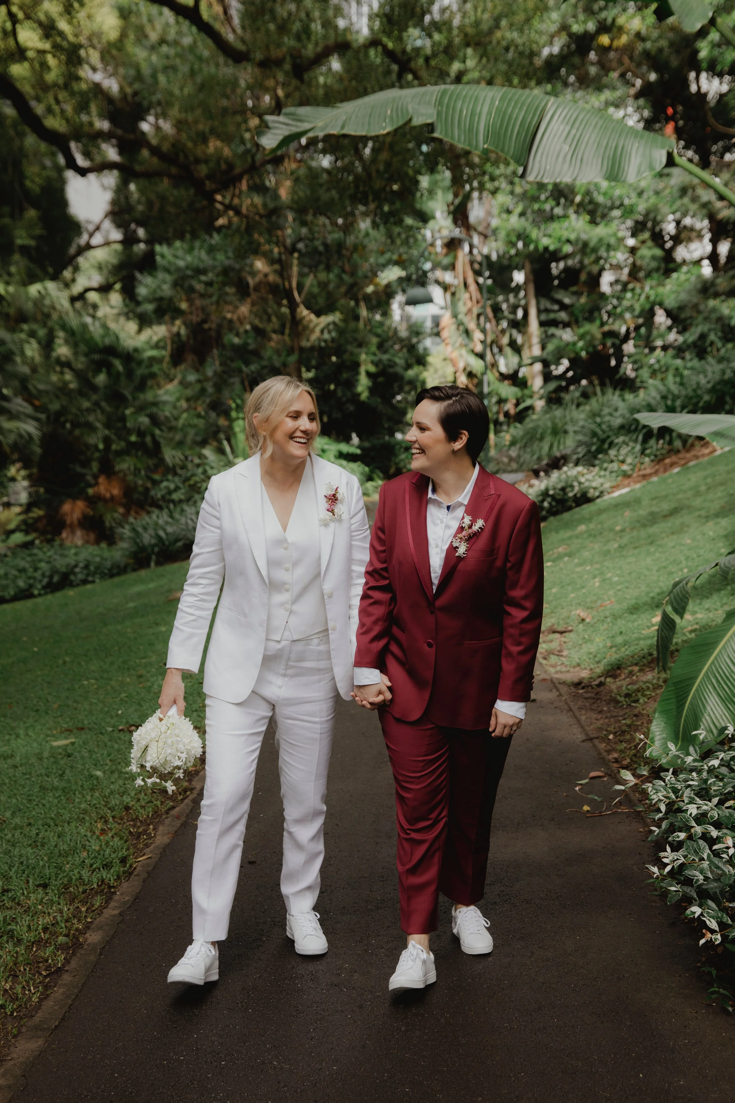 Two women in suits, one in white and one in maroon, holding hands and walking in a lush, green outdoor setting, smiling at each other.