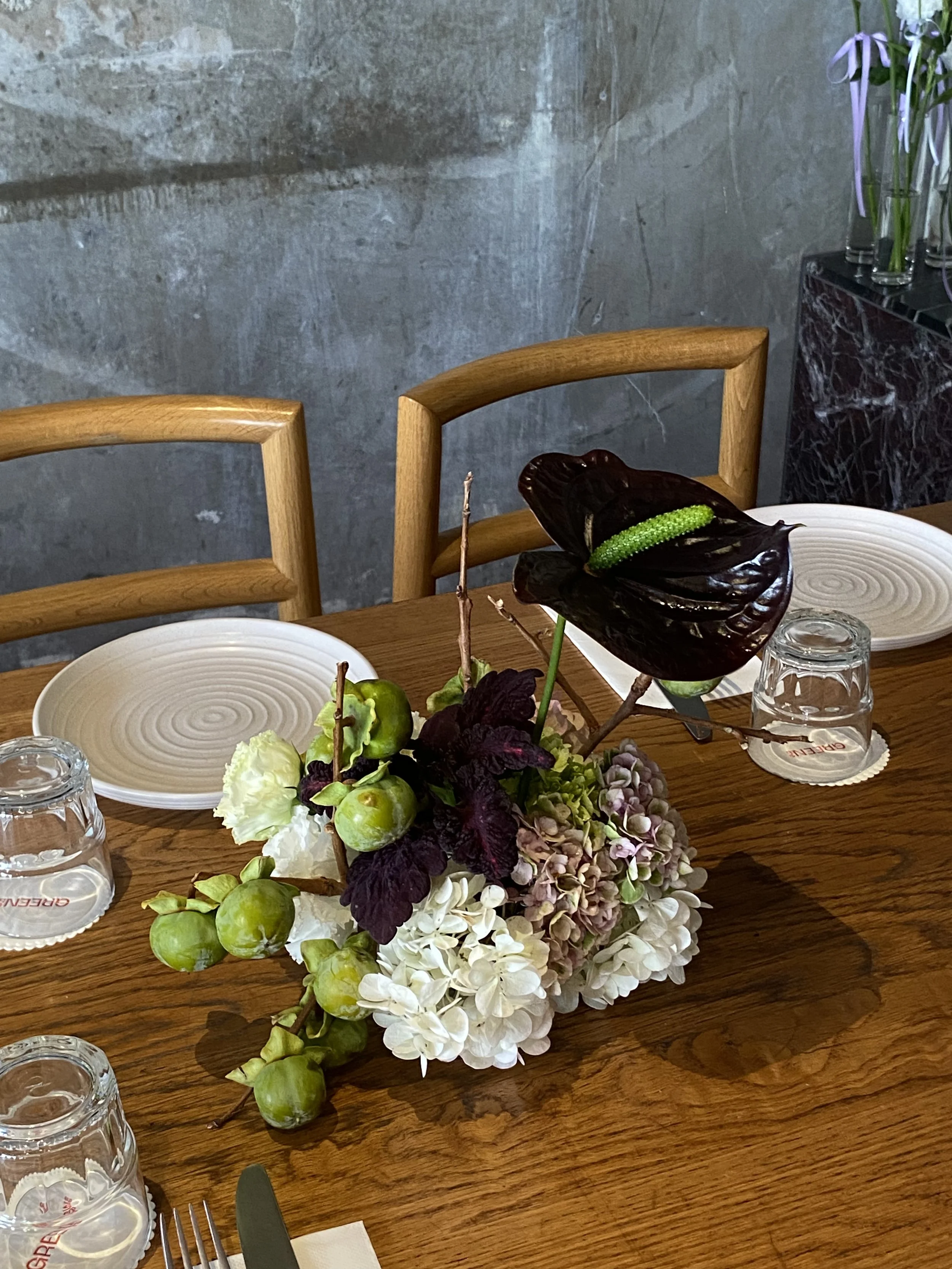 A wooden dining table arranged with a floral centerpiece, white plates, drinking glasses, and utensils, with chairs and a gray stone wall in the background.