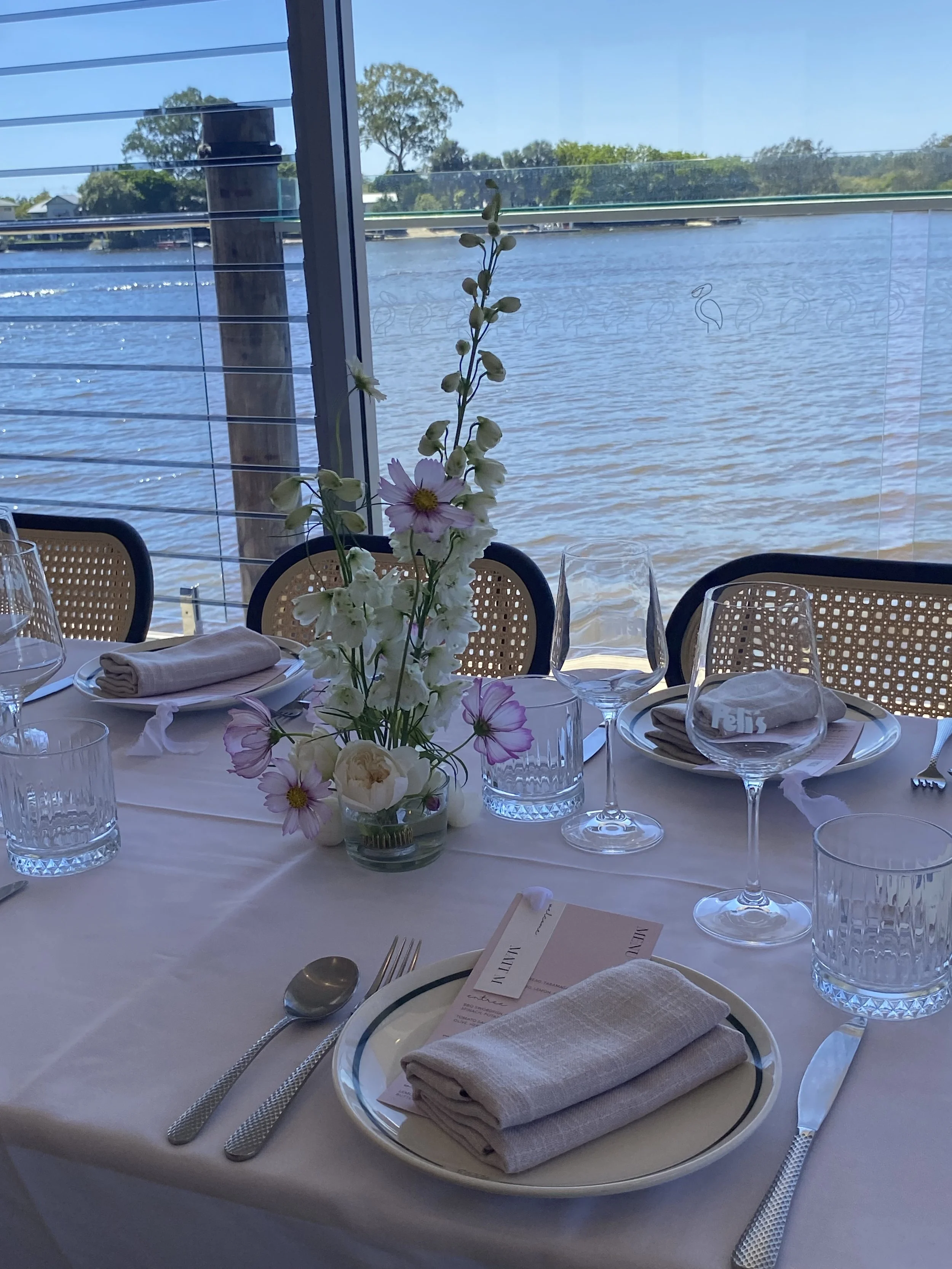 A dining table set with plates, napkins, wine glasses, and silverware, decorated with a tall floral centerpiece, overlooking a river outside with trees and a blue sky.