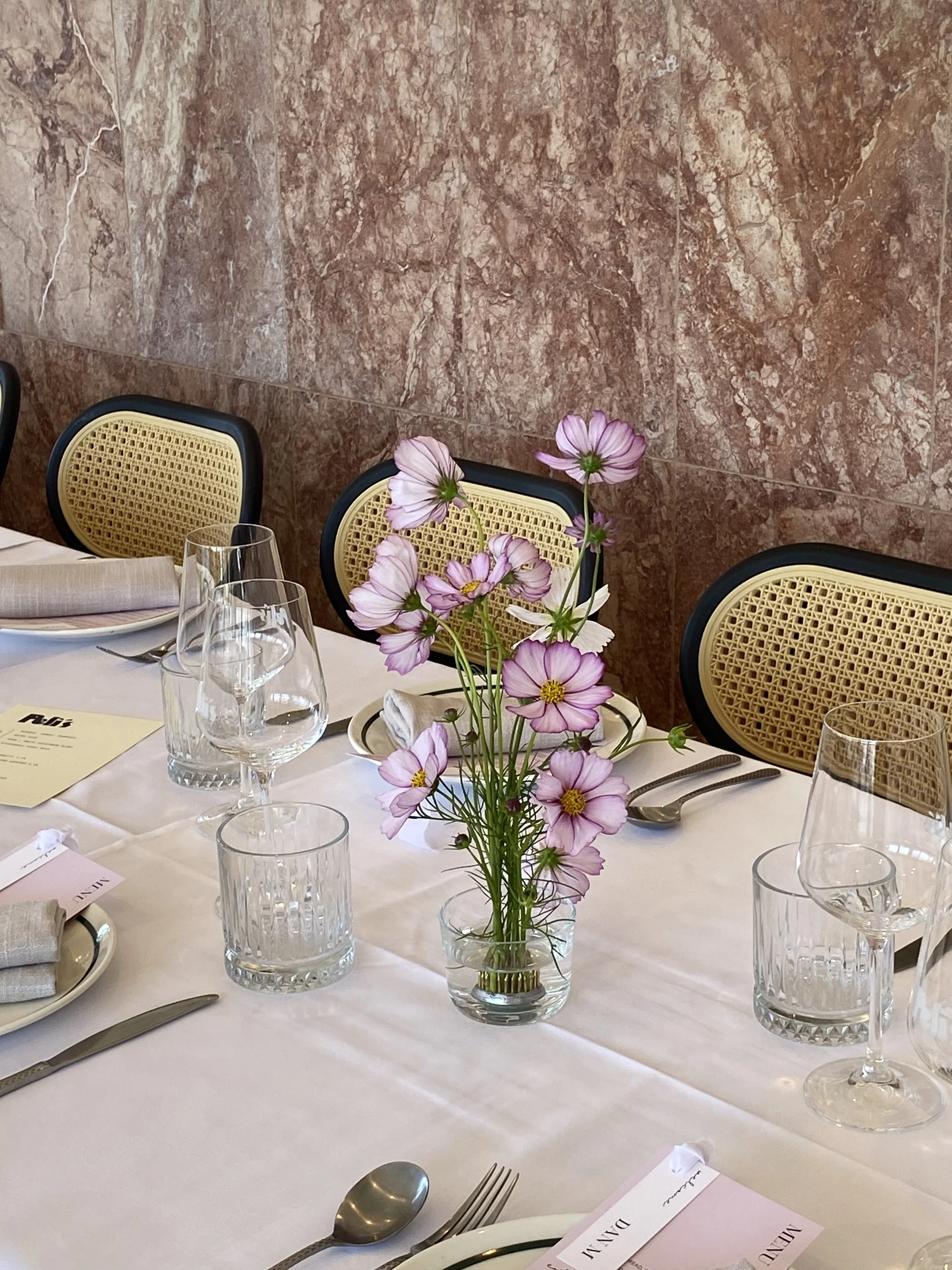 A dining table set with glasses, silverware, a menu, and a centerpiece of pink and white flowers in a clear glass vase, against a textured brown marble wall.