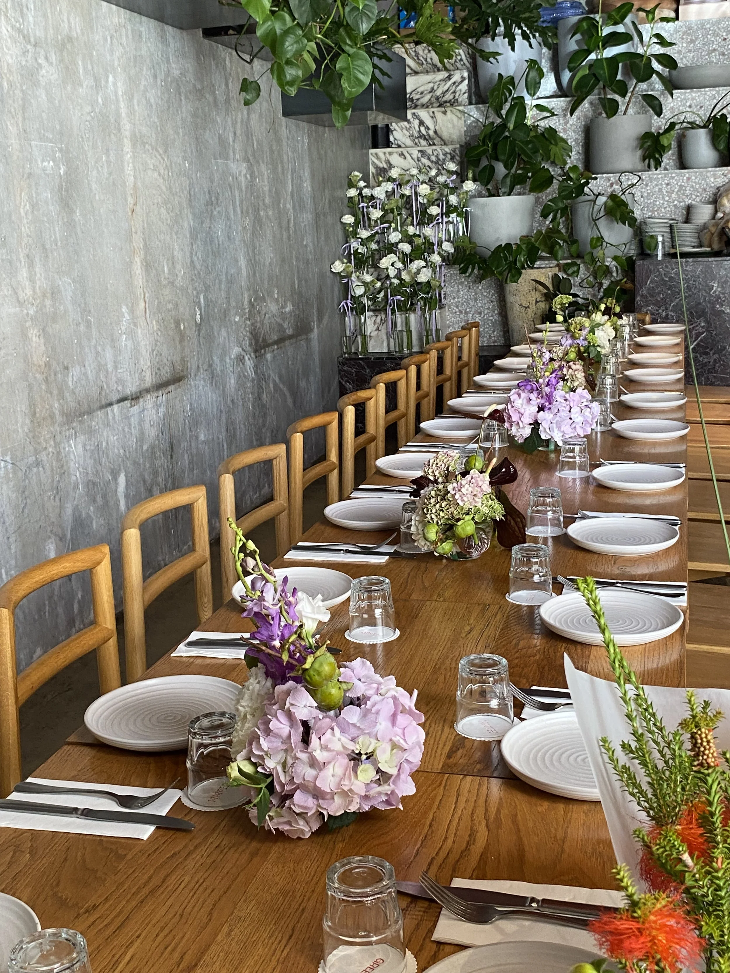 A long wooden dining table set for a formal event, decorated with pink and purple flowers in vases, white plates, glasses, and napkins, with wooden chairs around it. In the background, there are potted plants on shelves and a bunch of white and purpl
