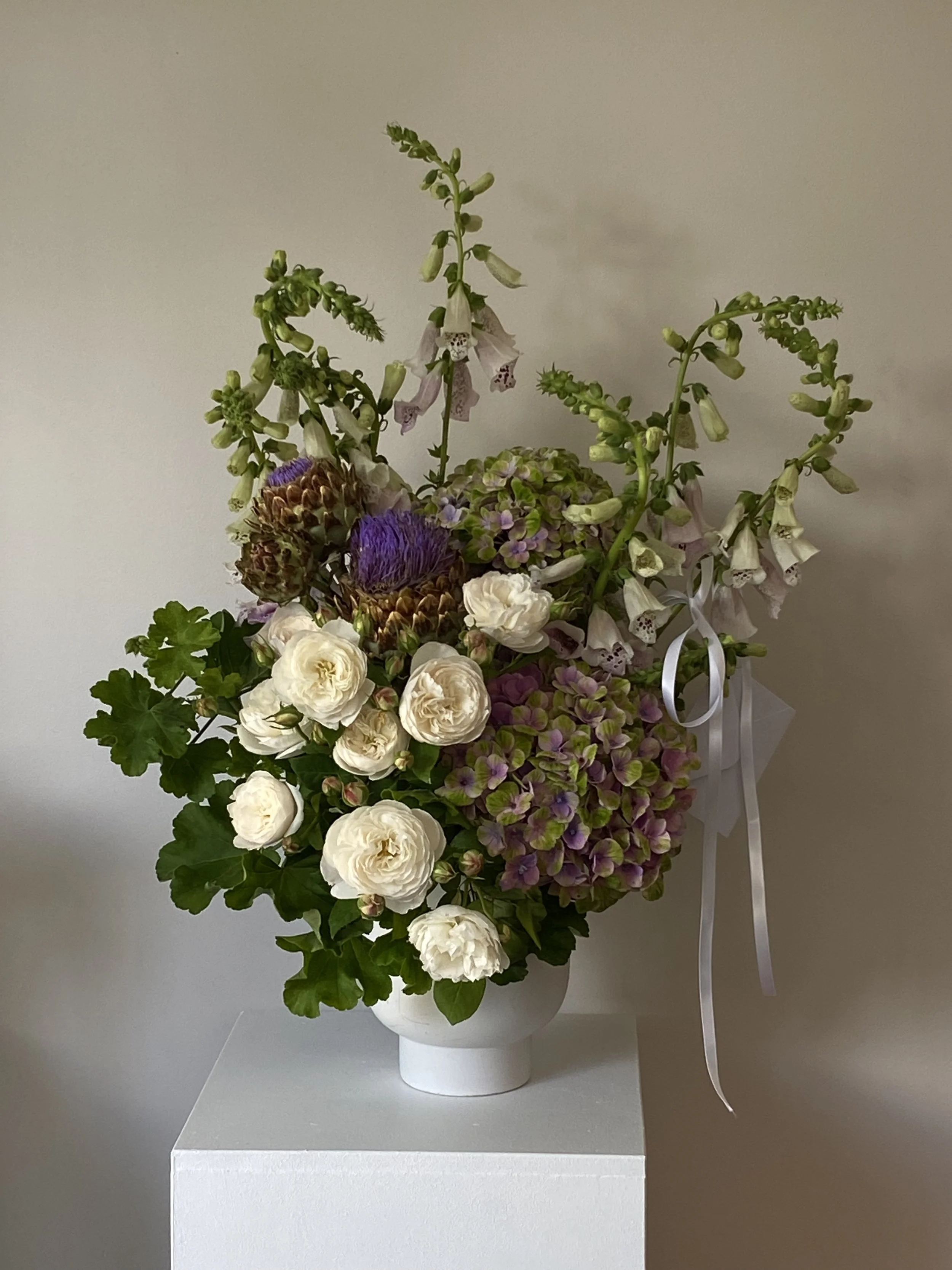 A tall floral arrangement in a white vase featuring white roses, purple thistle, hydrangeas in pink and purple, and green foliage, placed on a white pedestal against a plain wall.