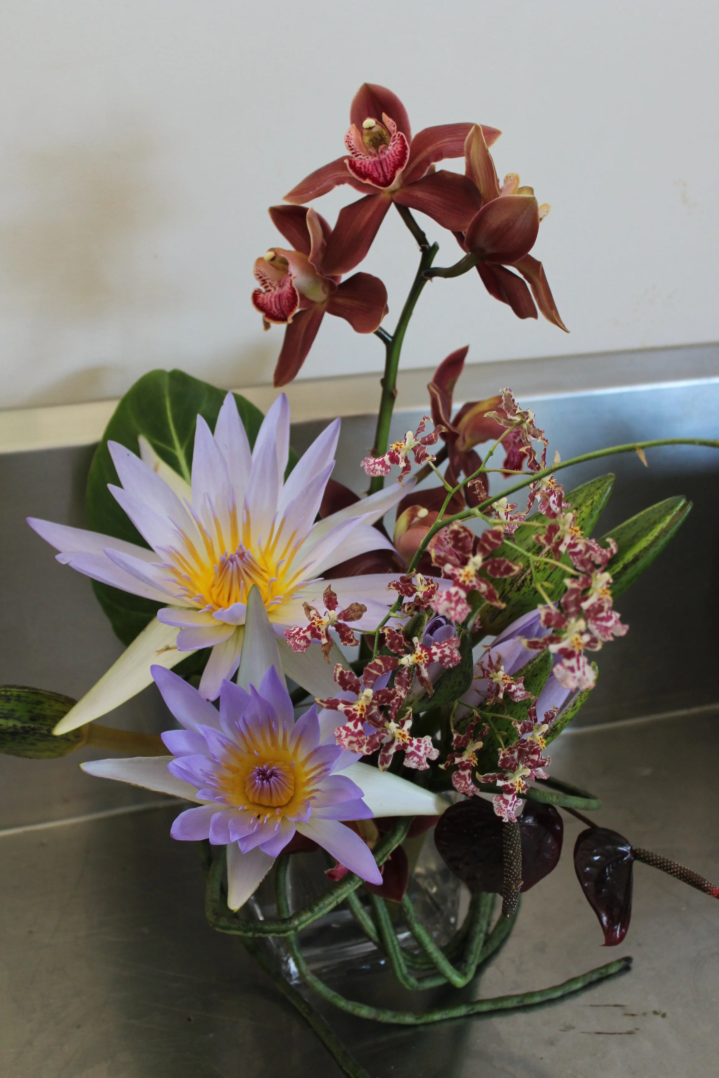 A floral arrangement featuring purple water lilies, red orchids, and greenery in a glass vase.